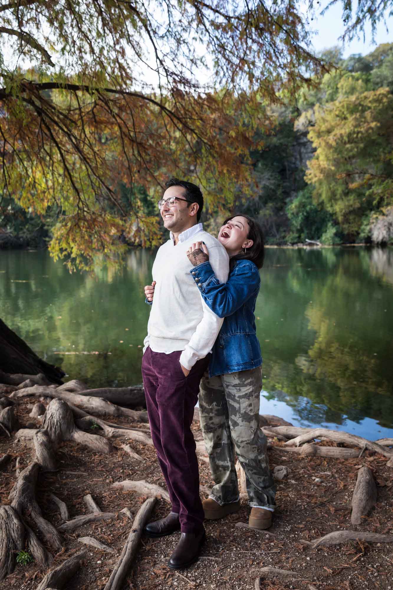Woman wearing denim jacket standing behind man wearing grey sweater hugging in front of river and tree with colorful leaves during a Red Bud Isle surprise proposal and engagement portrait