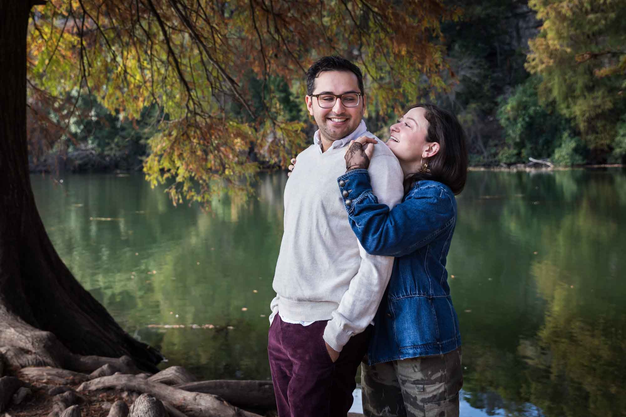 Woman wearing denim jacket standing behind man wearing grey sweater hugging in front of river and tree with colorful leaves during a Red Bud Isle surprise proposal and engagement portrait
