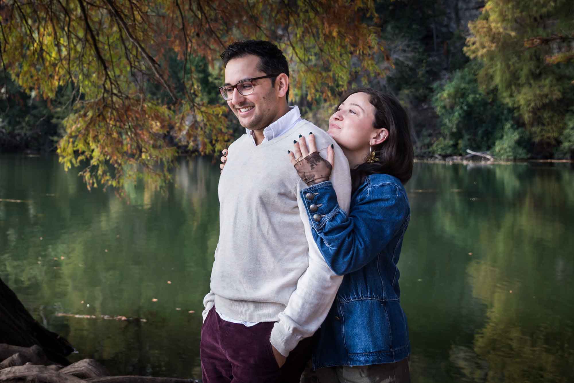 Woman wearing denim jacket standing behind man wearing grey sweater hugging in front of river and tree with colorful leaves during a Red Bud Isle surprise proposal and engagement portrait