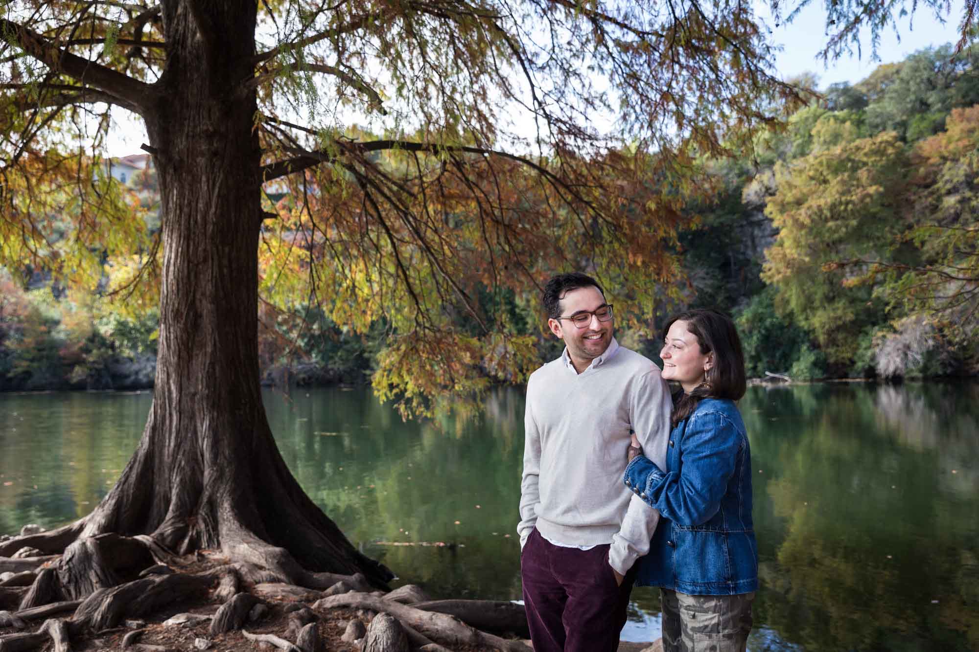 Woman wearing denim jacket standing behind man wearing grey sweater hugging in front of river and tree with colorful leaves during a Red Bud Isle surprise proposal and engagement portrait