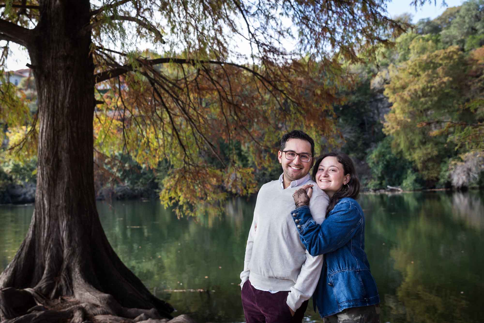 Woman wearing denim jacket standing behind man wearing grey sweater hugging in front of river and tree with colorful leaves during a Red Bud Isle surprise proposal and engagement portrait