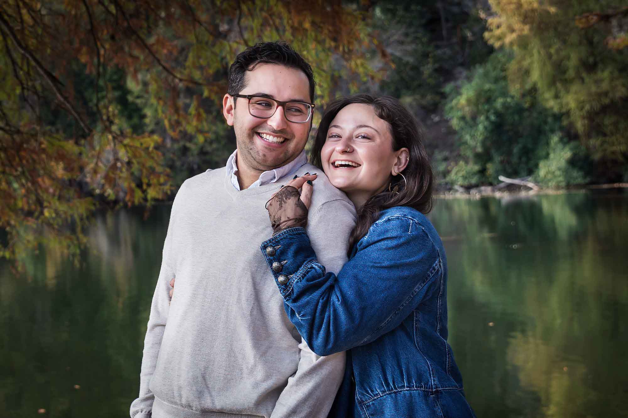 Woman wearing denim jacket standing behind man wearing grey sweater hugging in front of river and tree with colorful leaves during a Red Bud Isle surprise proposal and engagement portrait