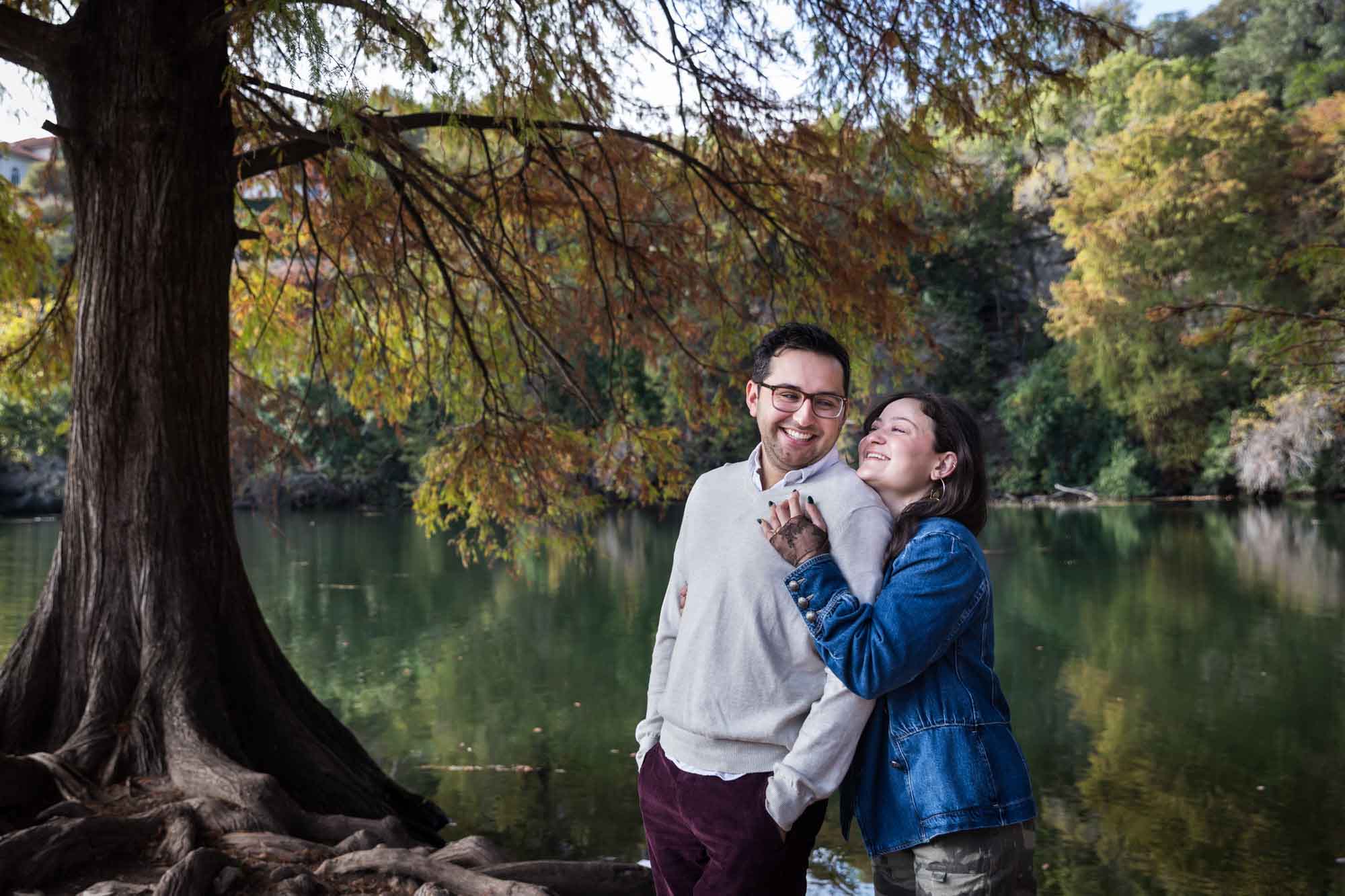Woman wearing denim jacket standing behind man wearing grey sweater hugging in front of river and tree with colorful leaves during a Red Bud Isle surprise proposal and engagement portrait