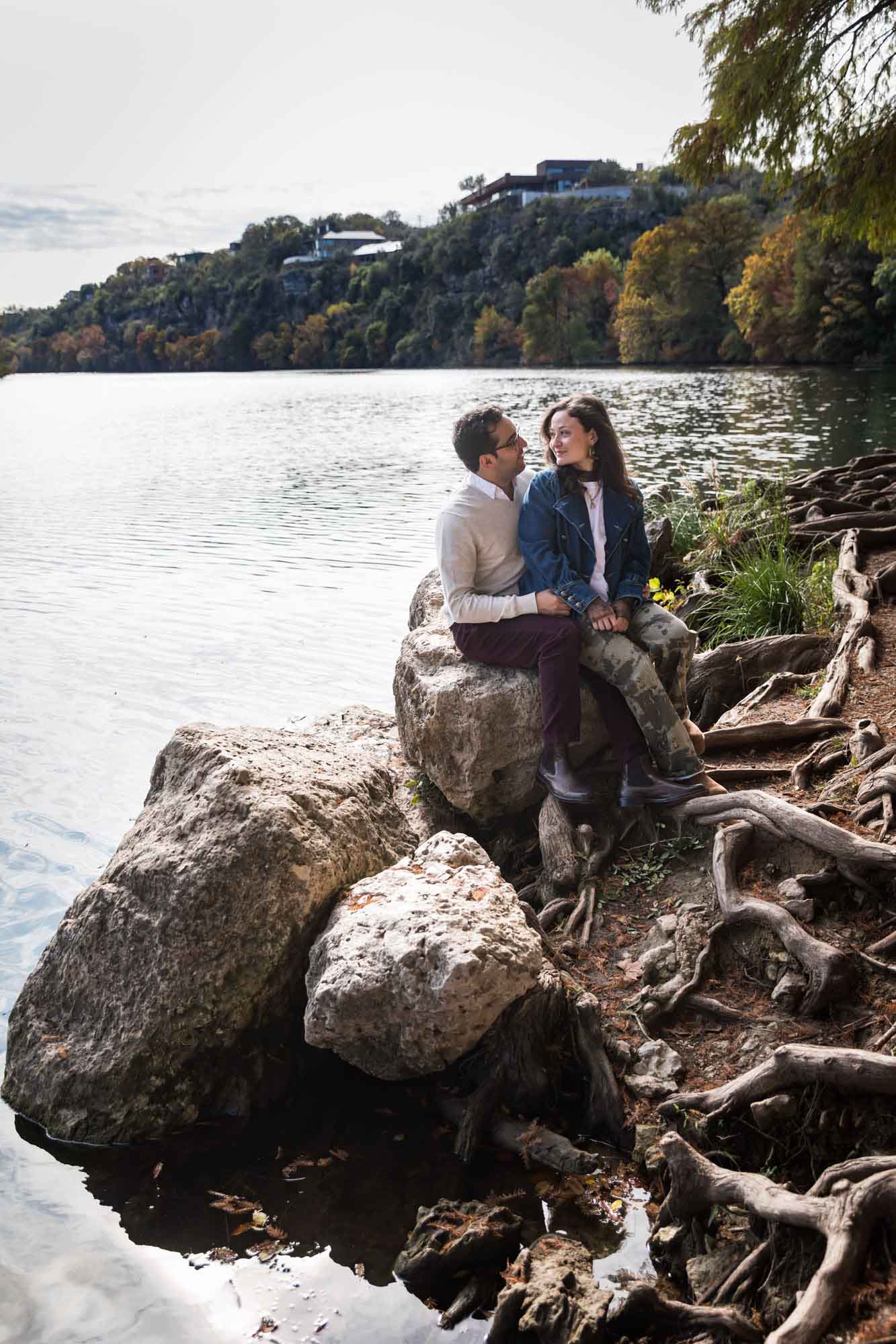 Man wearing grey sweater and maroon pants sitting on rock in front of river with woman wearing denim jacket and camo pants during a Red Bud Isle surprise proposal