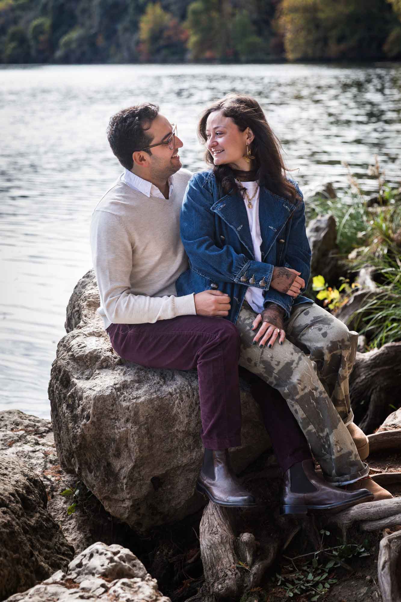 Man wearing grey sweater and maroon pants sitting on rock in front of river with woman wearing denim jacket and camo pants during a Red Bud Isle surprise proposal