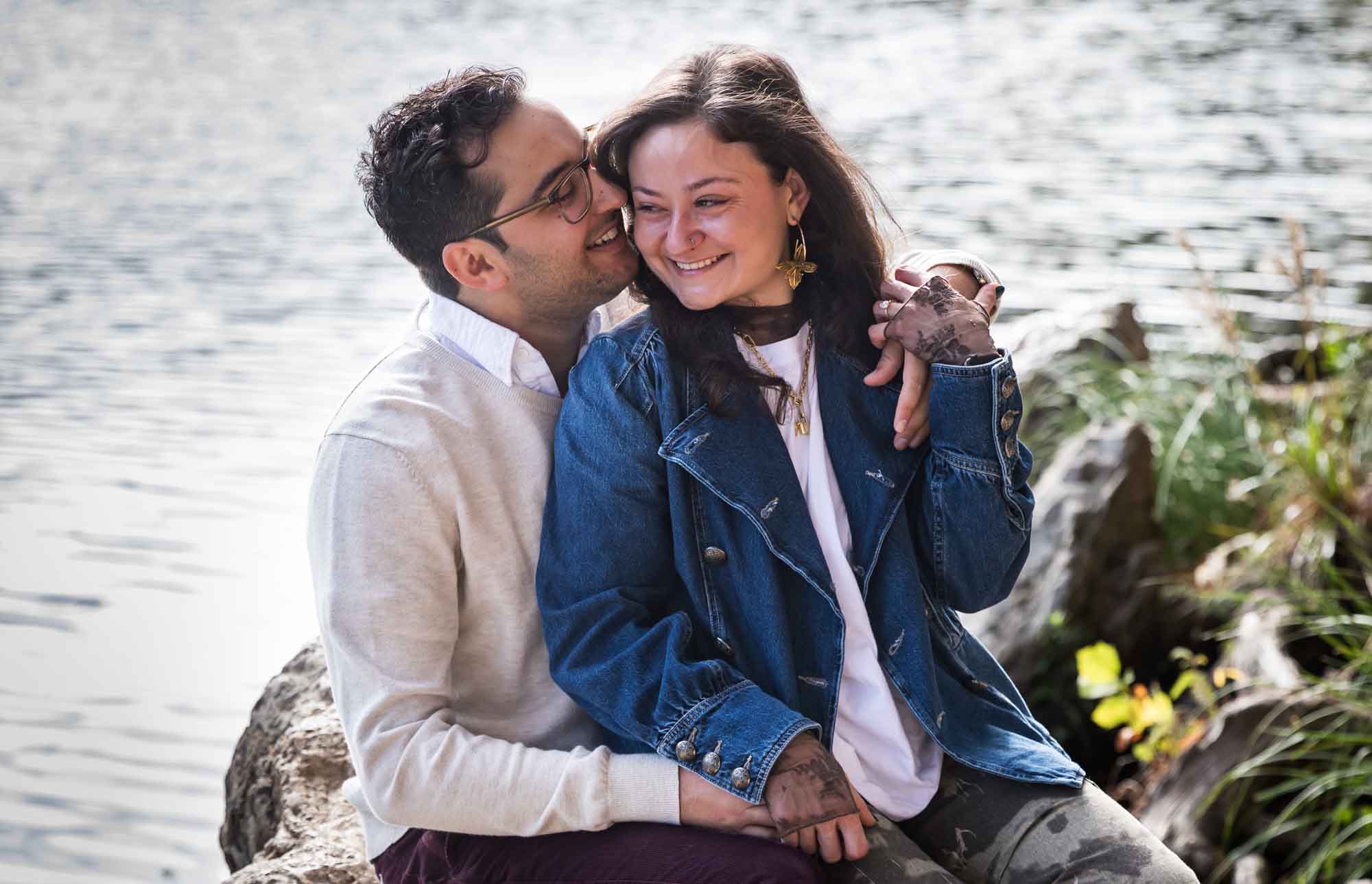 Man wearing grey sweater and maroon pants sitting on rock in front of river with woman wearing denim jacket and camo pants during a Red Bud Isle surprise proposal