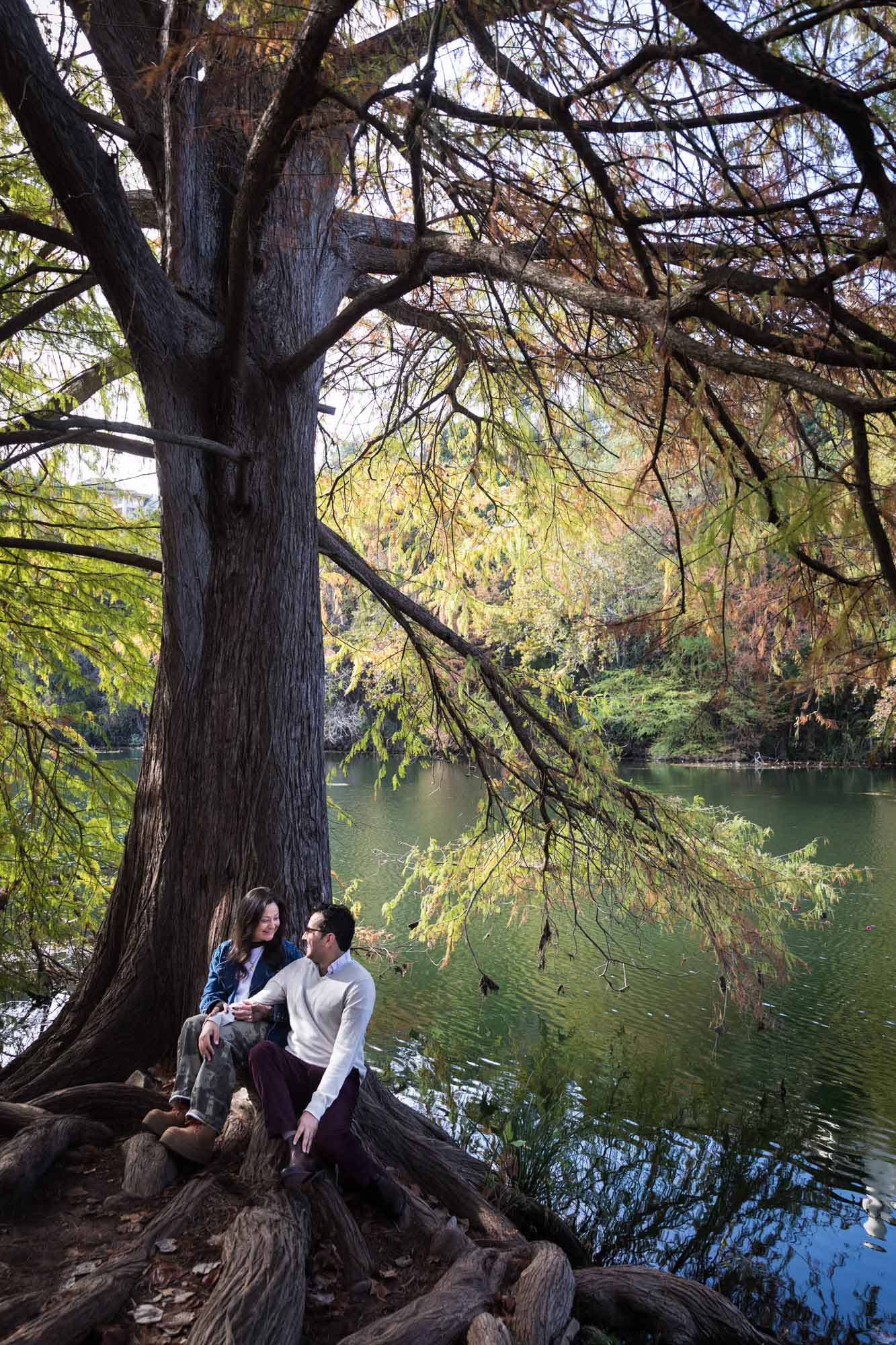 Man wearing grey sweater and maroon pants sitting on large tree root in front of river with woman wearing denim jacket and camo pants during a Red Bud Isle surprise proposal