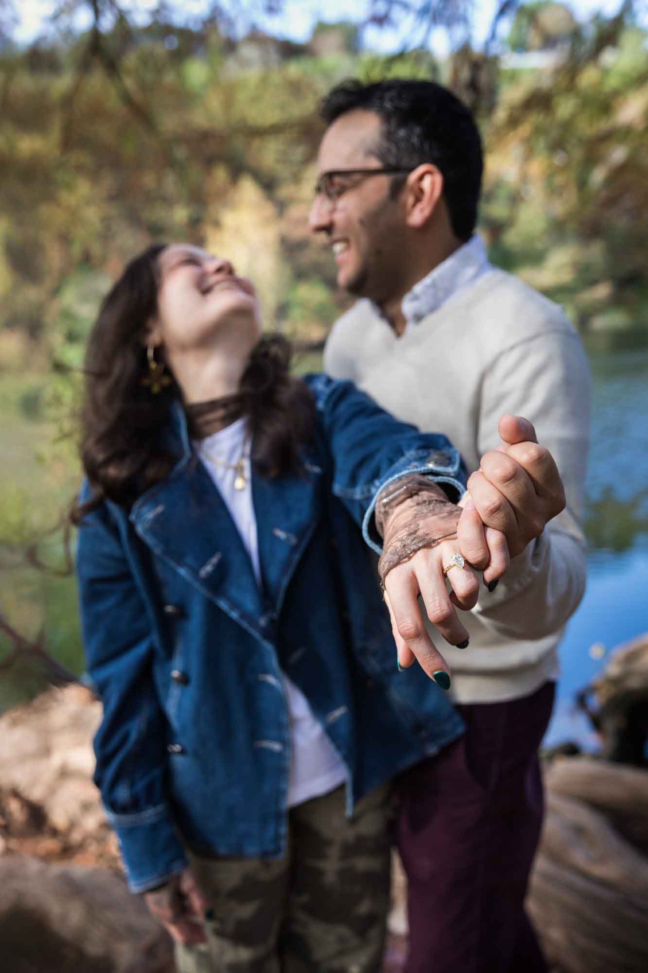 Woman wearing denim jacket laughing with man wearing grey sweater showing her hand to camera with engagement ring