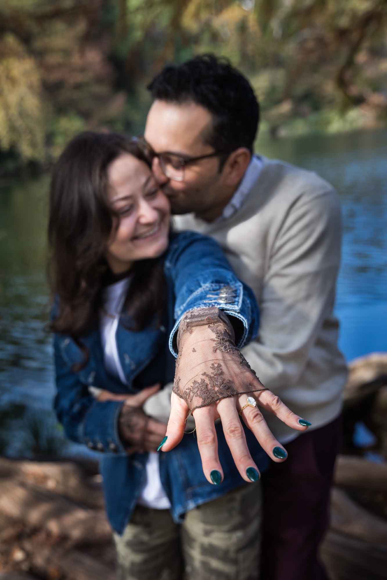Woman wearing denim jacket being kissed by man wearing grey sweater showing her hand to camera with engagement ring