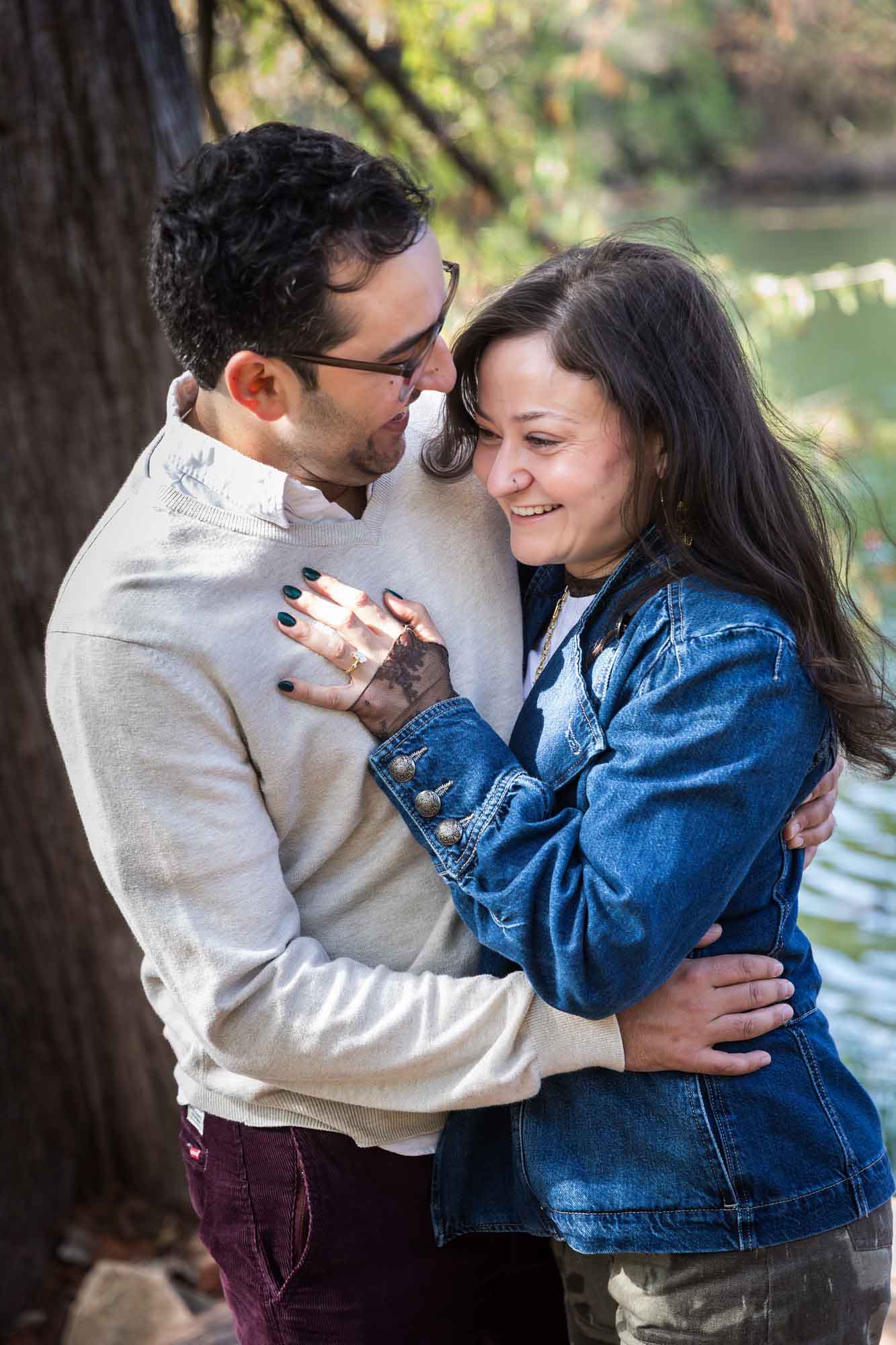 Woman wearing denim jacket hugging man wearing grey sweater in front of tree and river during a Red Bud Isle surprise proposal