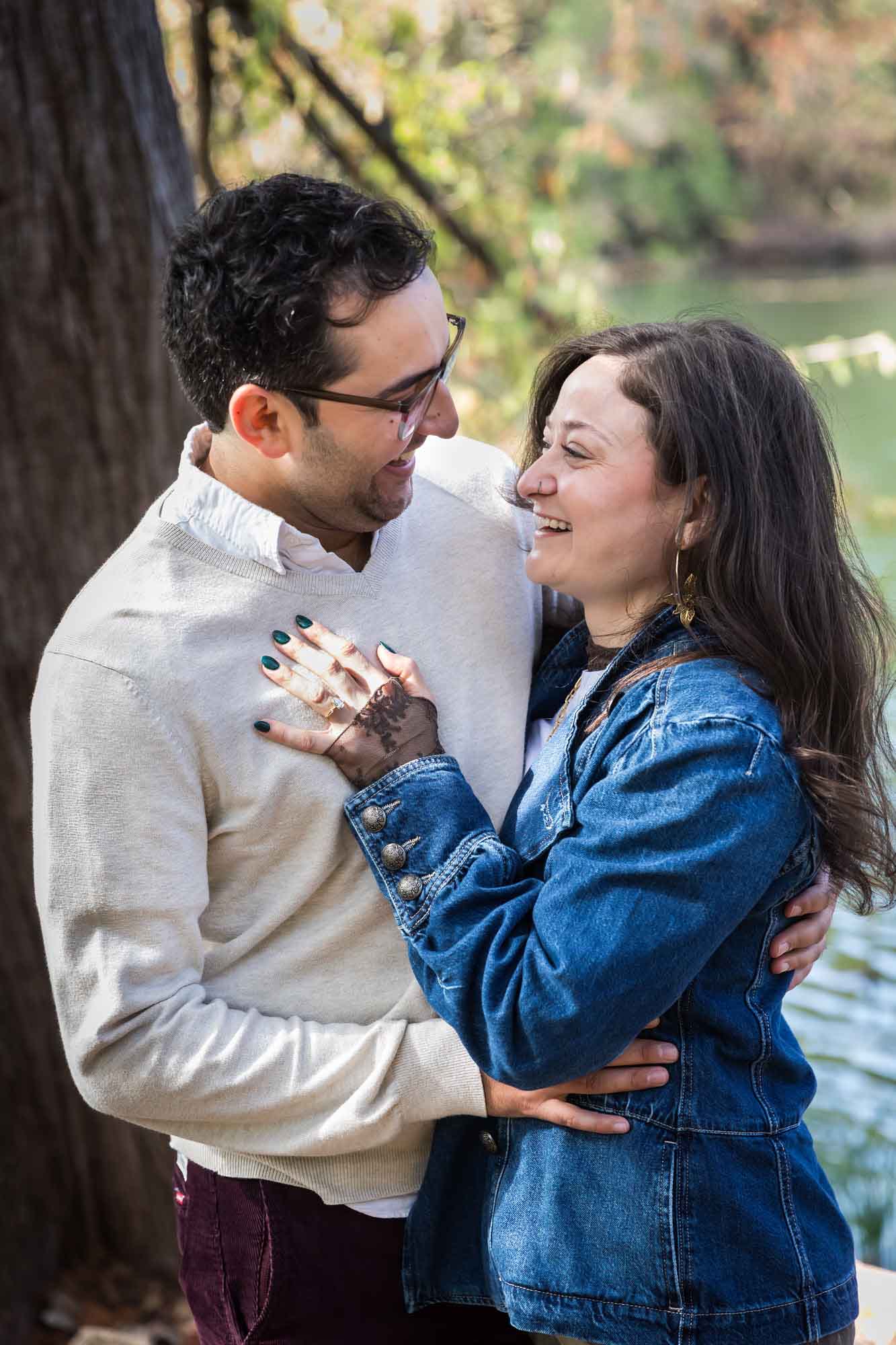 Woman wearing denim jacket hugging man wearing grey sweater in front of tree and river during a Red Bud Isle surprise proposal