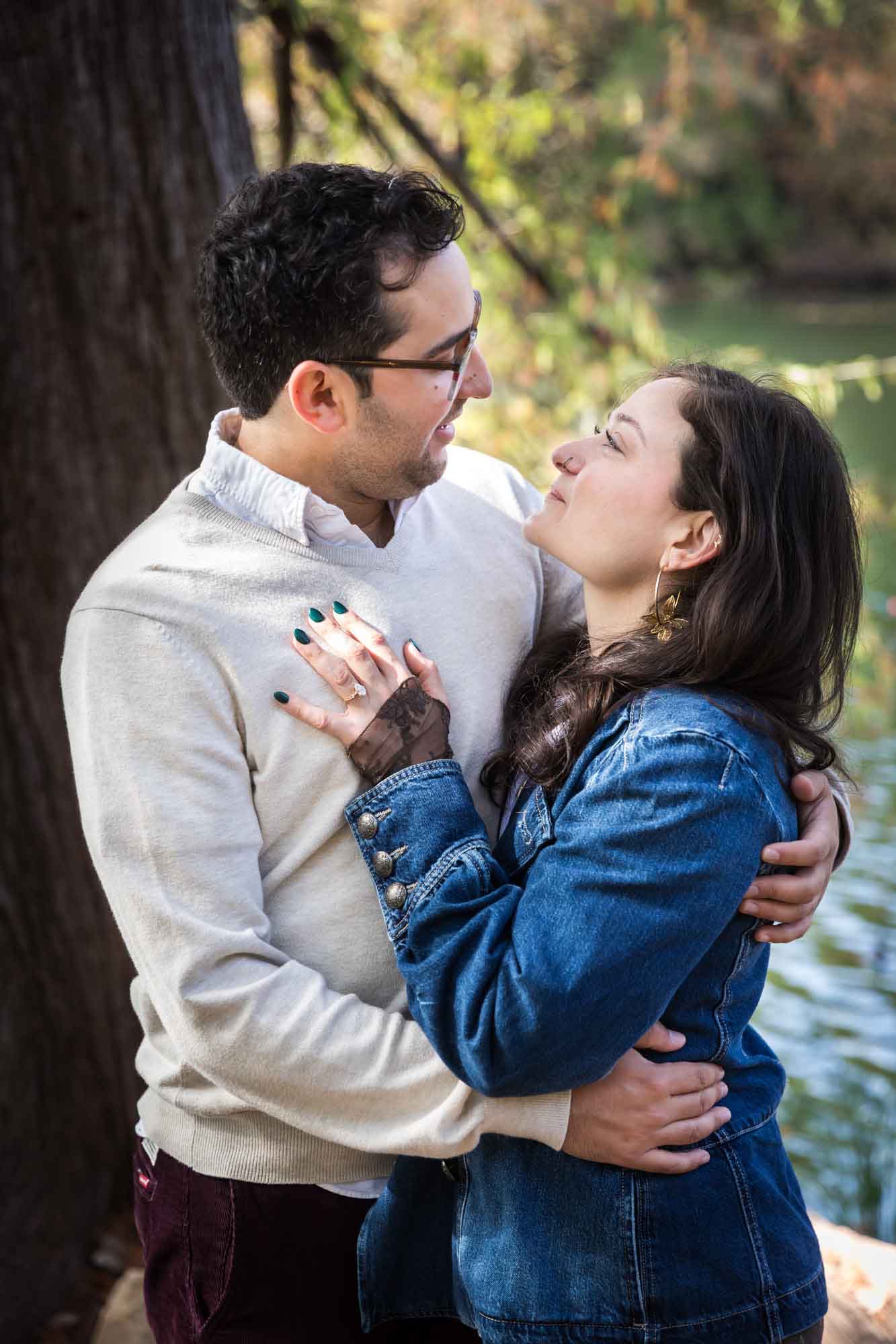 Woman wearing denim jacket hugging man wearing grey sweater in front of tree and river during a Red Bud Isle surprise proposal