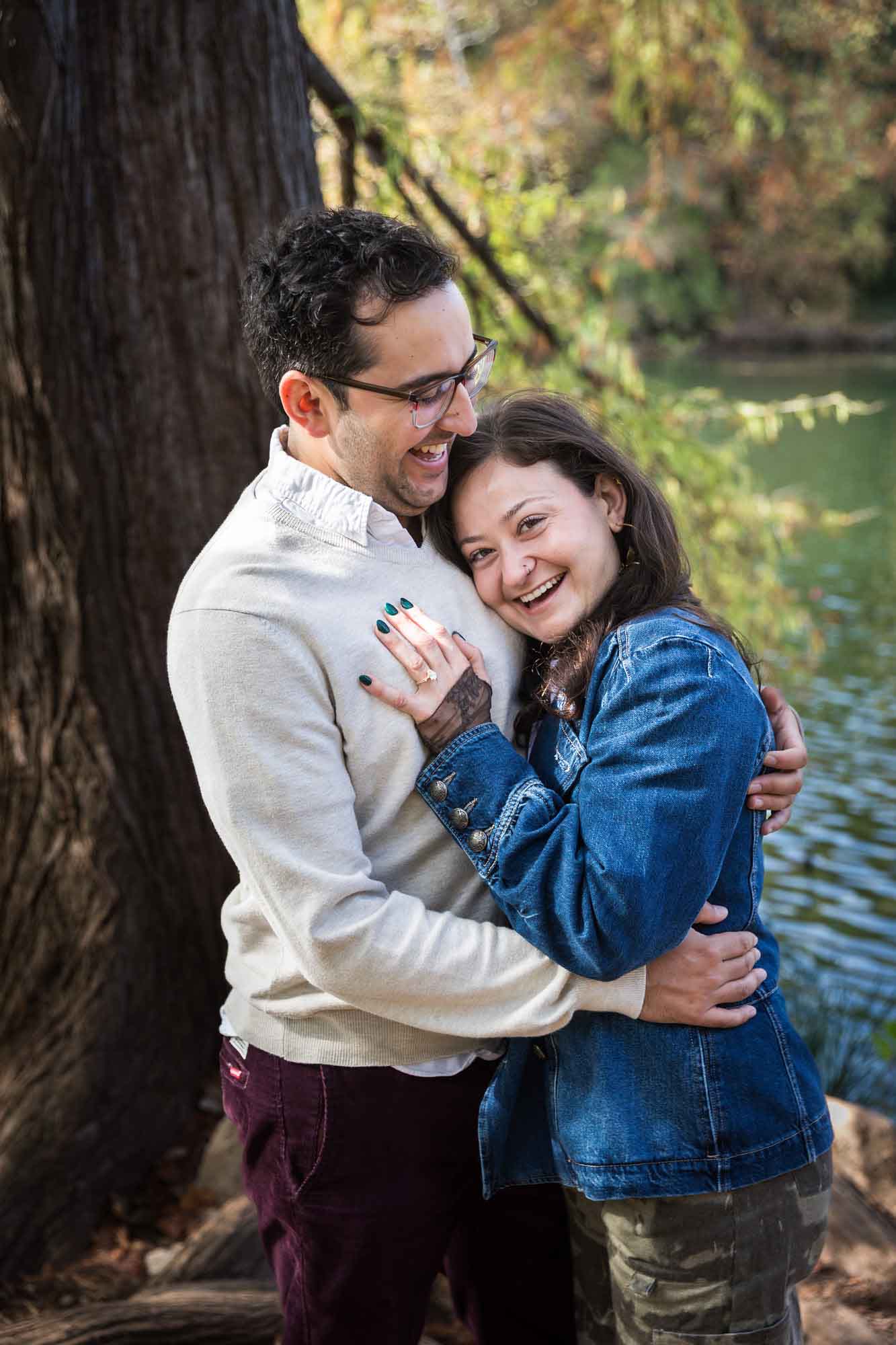 Woman wearing denim jacket hugging man wearing grey sweater in front of tree and river during a Red Bud Isle surprise proposal