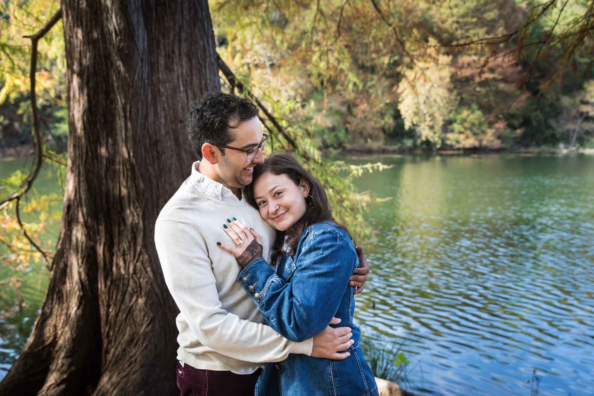Woman wearing denim jacket hugging man wearing grey sweater in front of tree and river during a Red Bud Isle surprise proposal