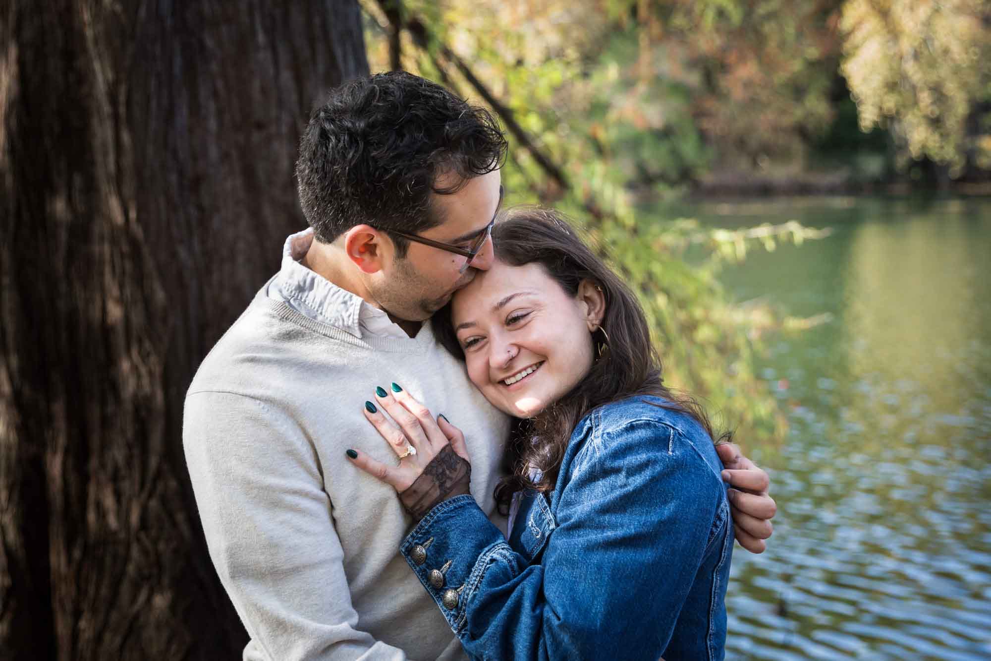 Woman wearing denim jacket hugging man wearing grey sweater in front of tree and river during a Red Bud Isle surprise proposal