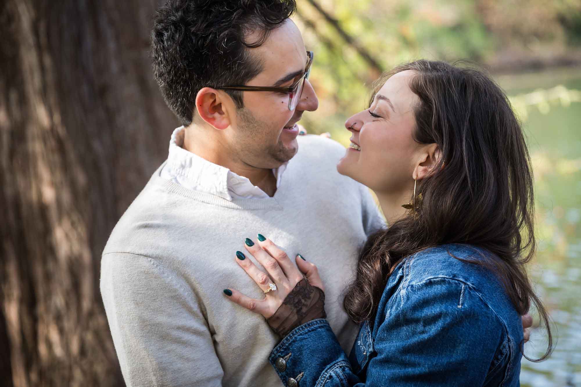 Woman wearing denim jacket hugging man wearing grey sweater in front of tree and river during a Red Bud Isle surprise proposal