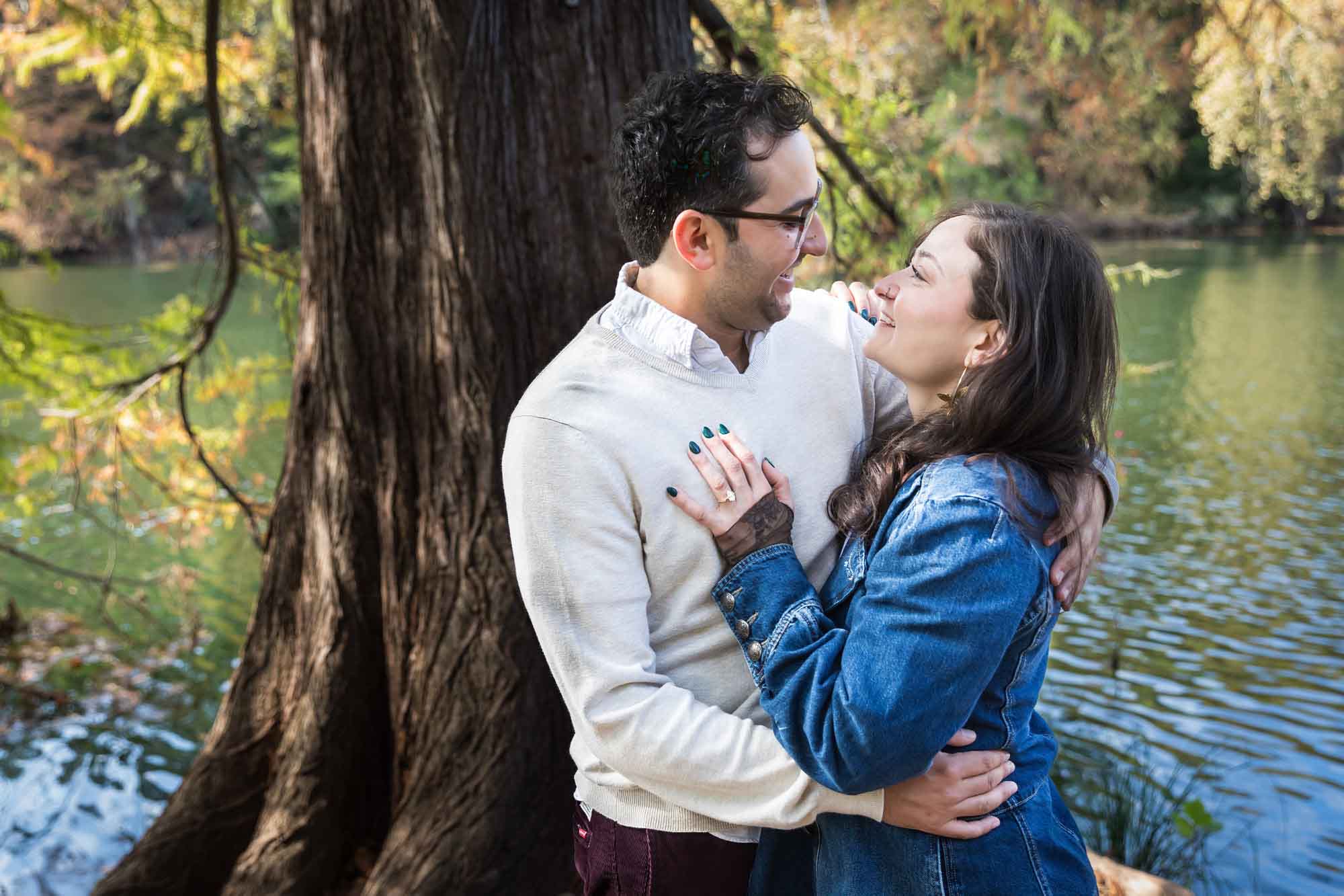 Woman wearing denim jacket hugging man wearing grey sweater in front of tree and river during a Red Bud Isle surprise proposal