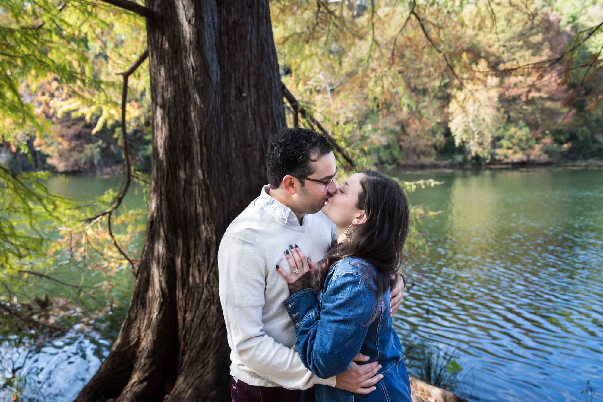 Woman wearing denim jacket kissing man wearing grey sweater in front of tree and river during a Red Bud Isle surprise proposal
