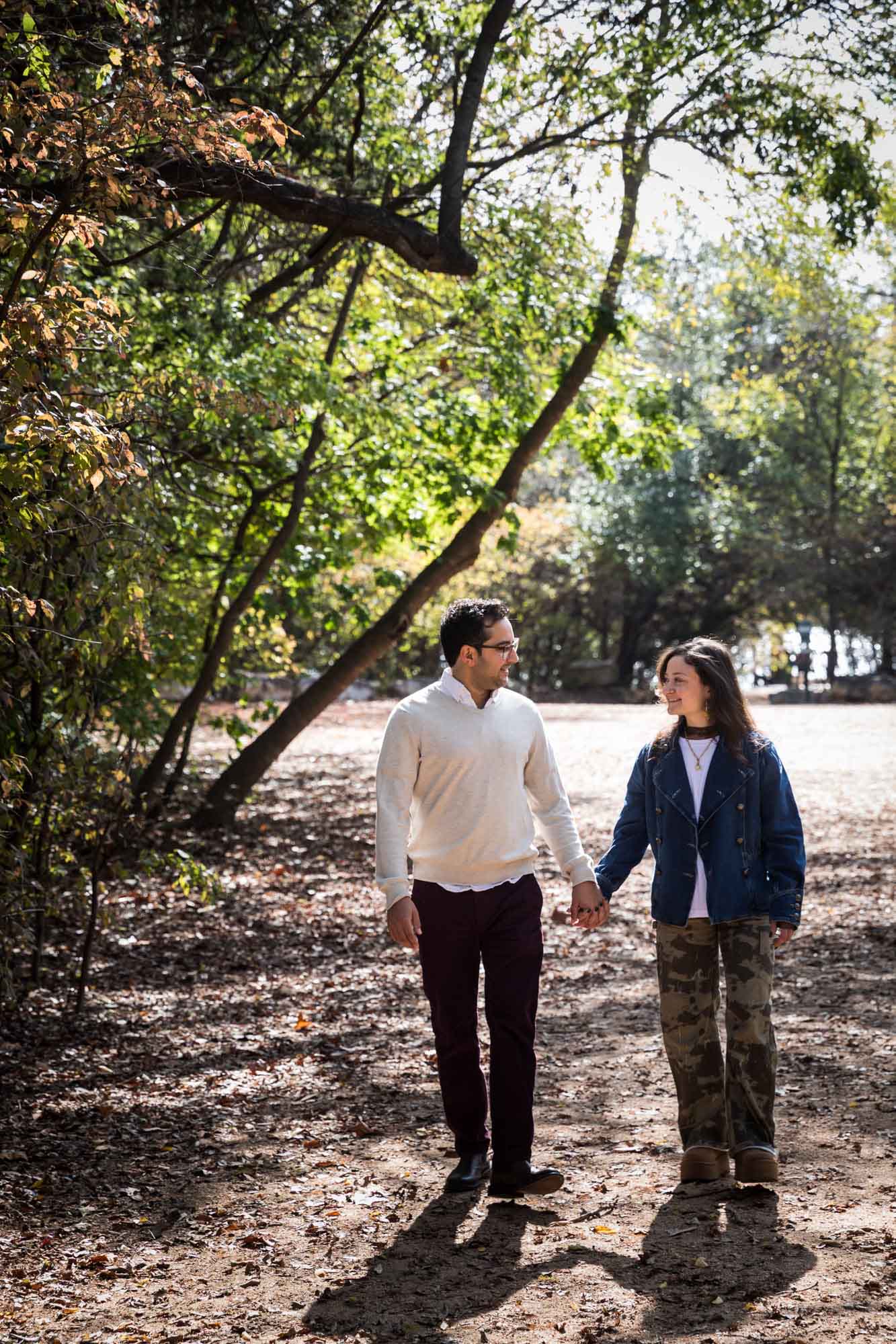 Woman wearing denim jacket holding hands and walking with man wearing grey sweater down forest pathway during a Red Bud Isle surprise proposal