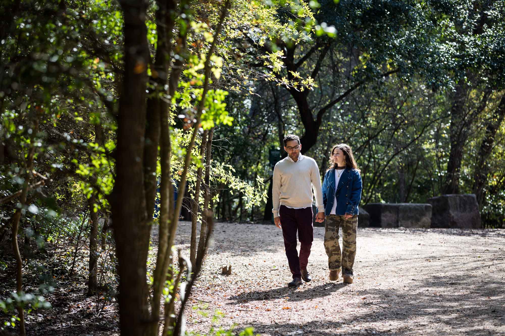 Woman wearing denim jacket holding hands and walking with man wearing grey sweater down forest pathway during a Red Bud Isle surprise proposal