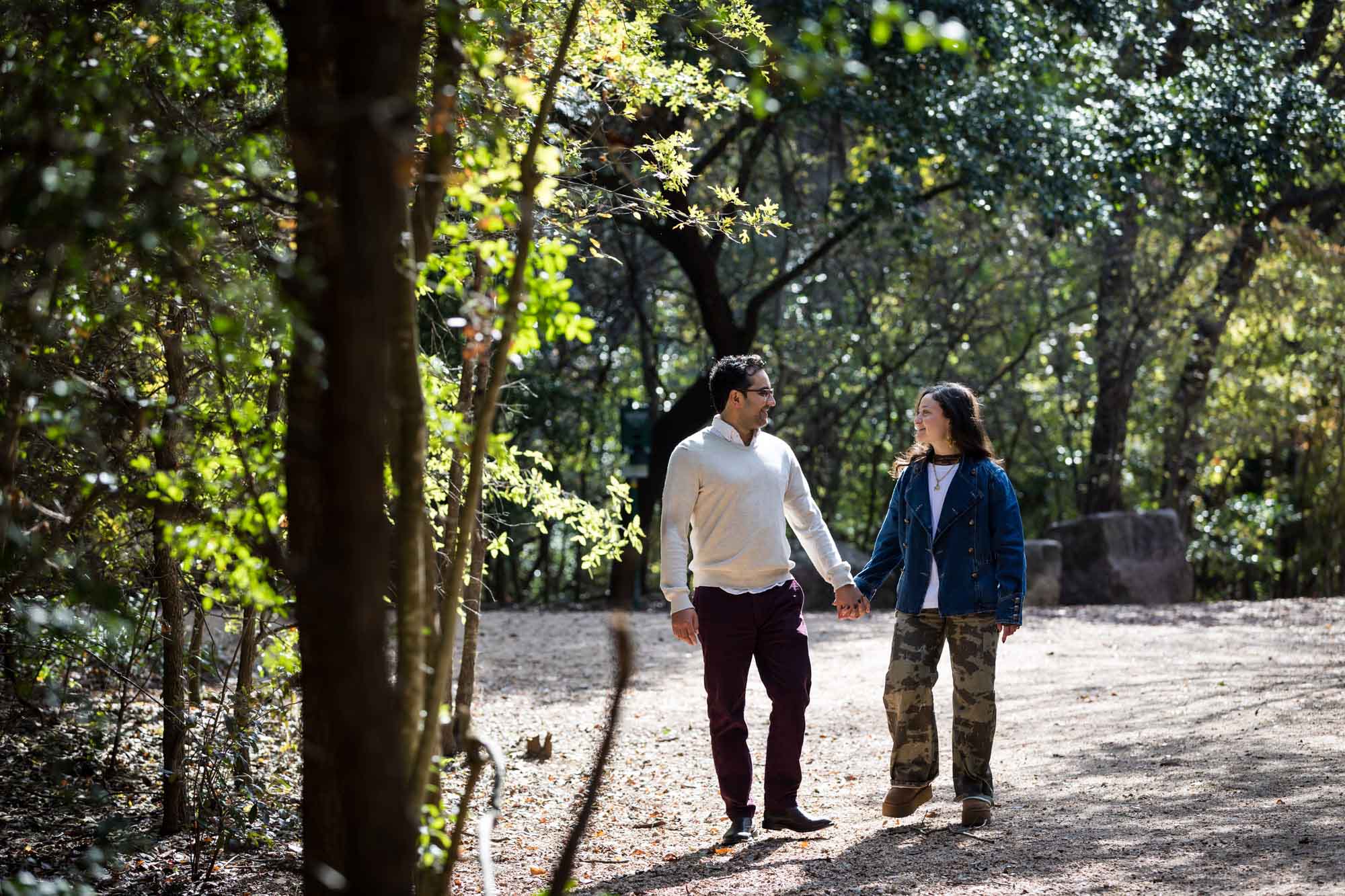 Woman wearing denim jacket holding hands and walking with man wearing grey sweater down forest pathway during a Red Bud Isle surprise proposal