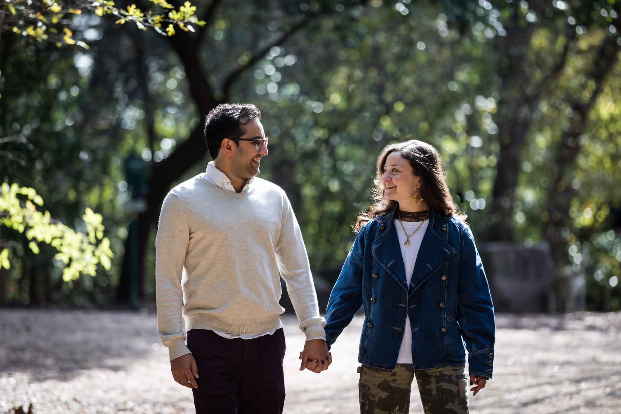 Woman wearing denim jacket holding hands and walking with man wearing grey sweater down forest pathway during a Red Bud Isle surprise proposal