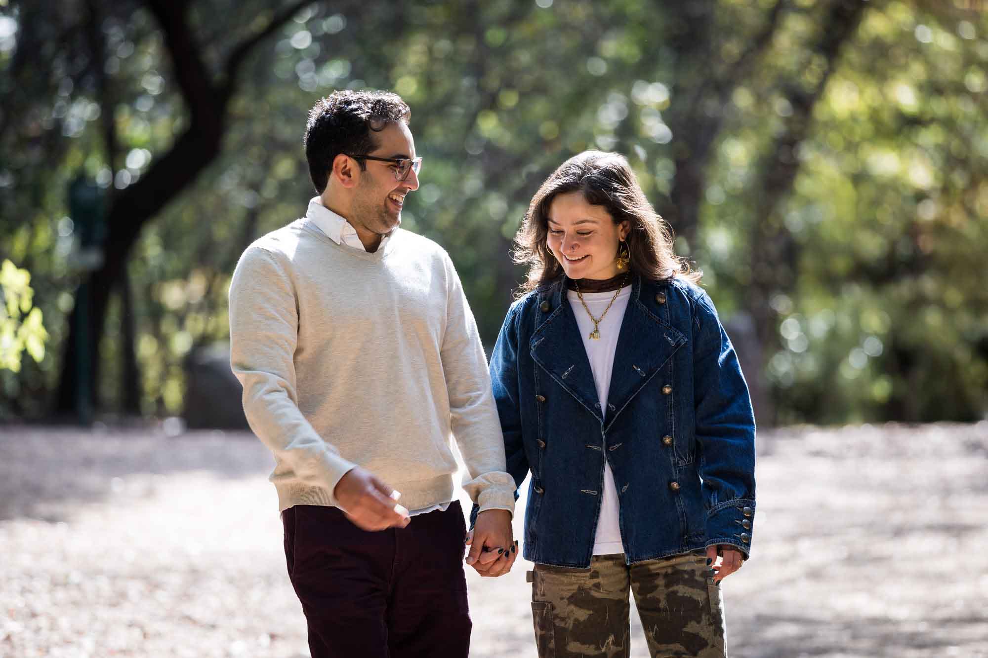 Woman wearing denim jacket holding hands and walking with man wearing grey sweater down forest pathway during a Red Bud Isle surprise proposal