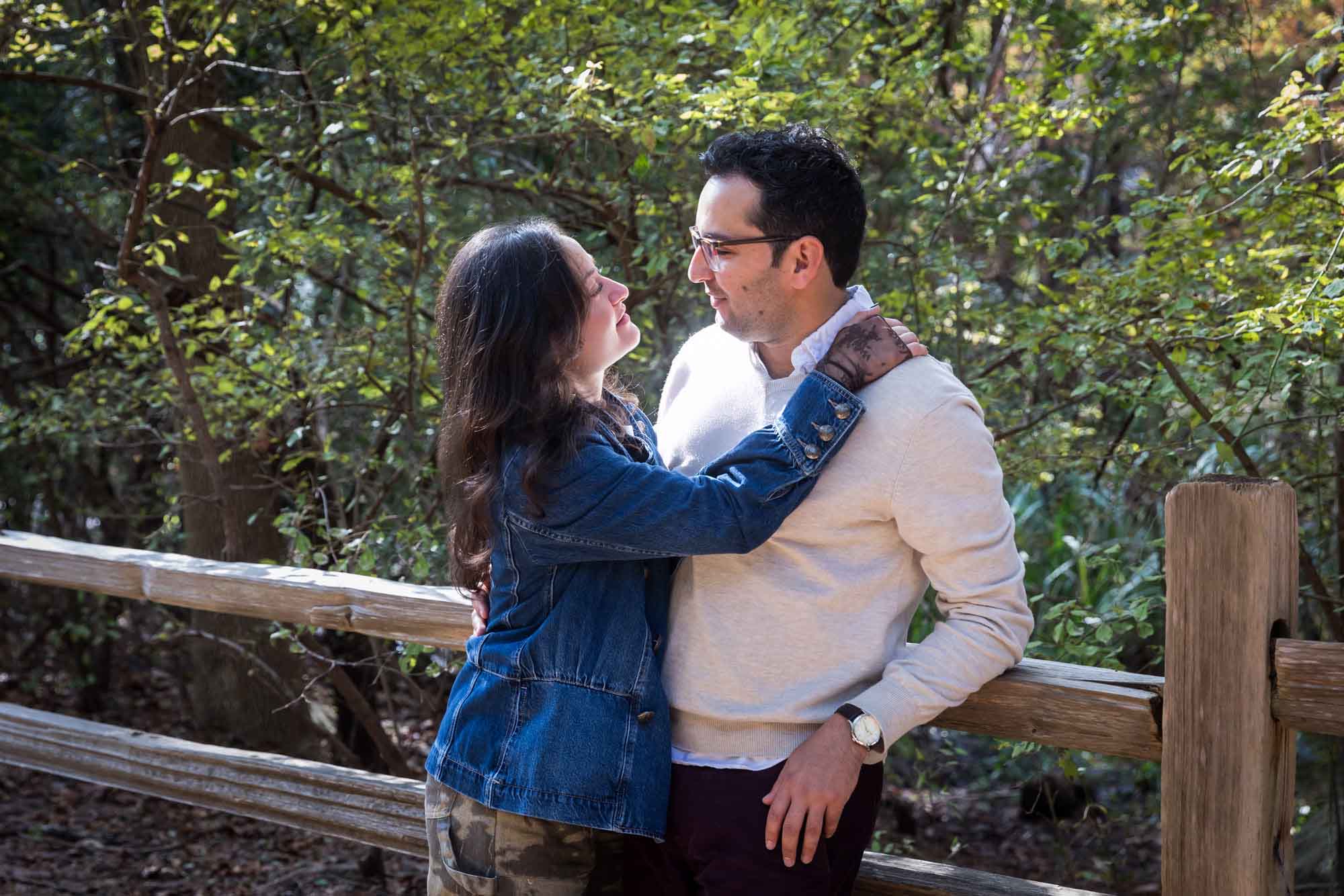 Woman wearing denim jacket hugging man wearing grey sweater against wooden railing in front of trees during a Red Bud Isle surprise proposal