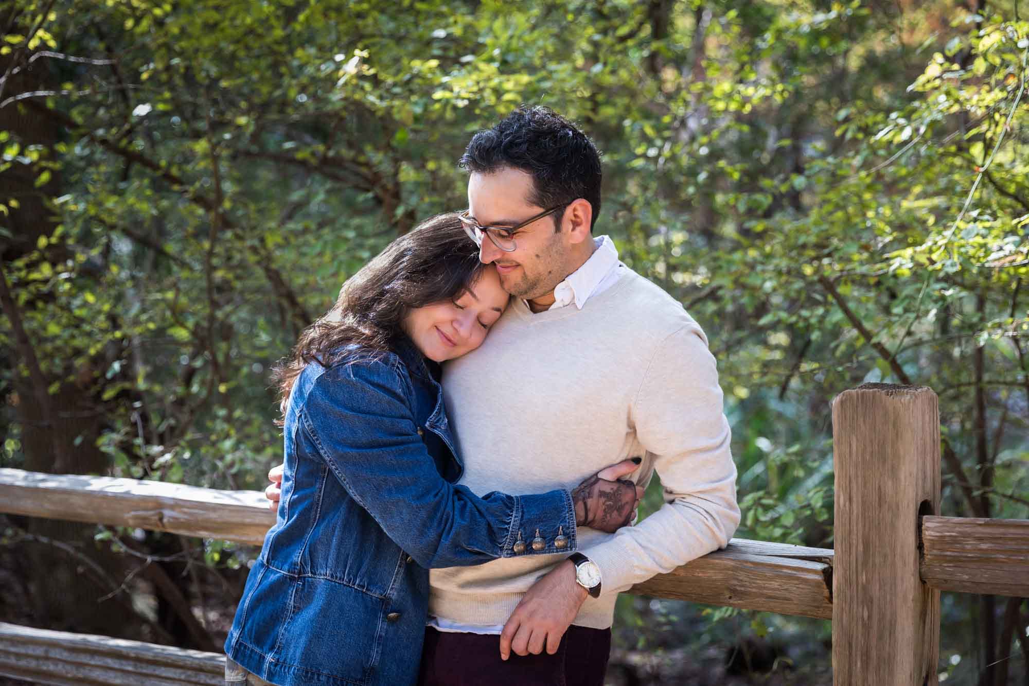 Woman wearing denim jacket hugging man wearing grey sweater against wooden railing in front of trees during a Red Bud Isle surprise proposal