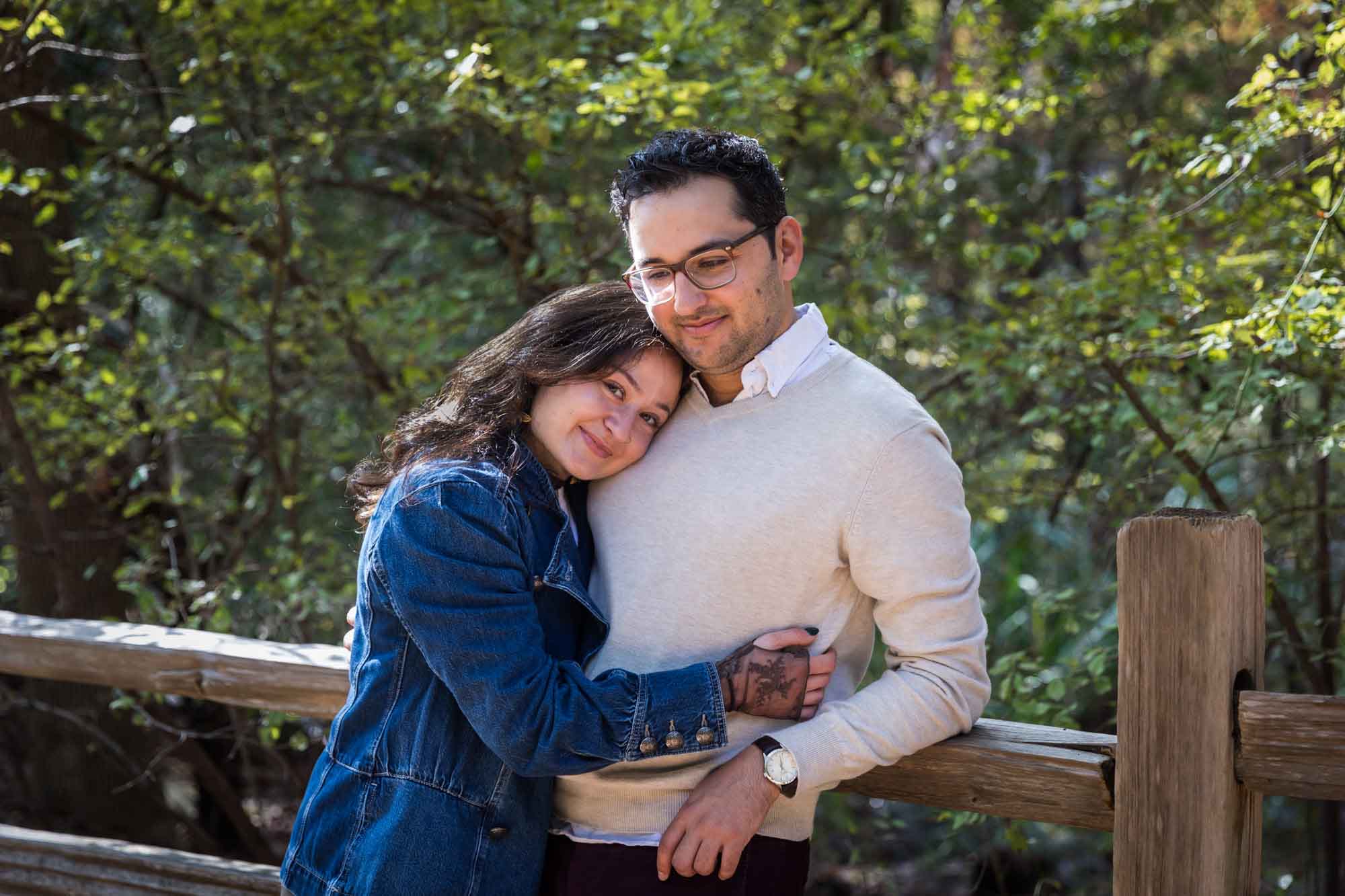 Woman wearing denim jacket hugging man wearing grey sweater against wooden railing in front of trees during a Red Bud Isle surprise proposal