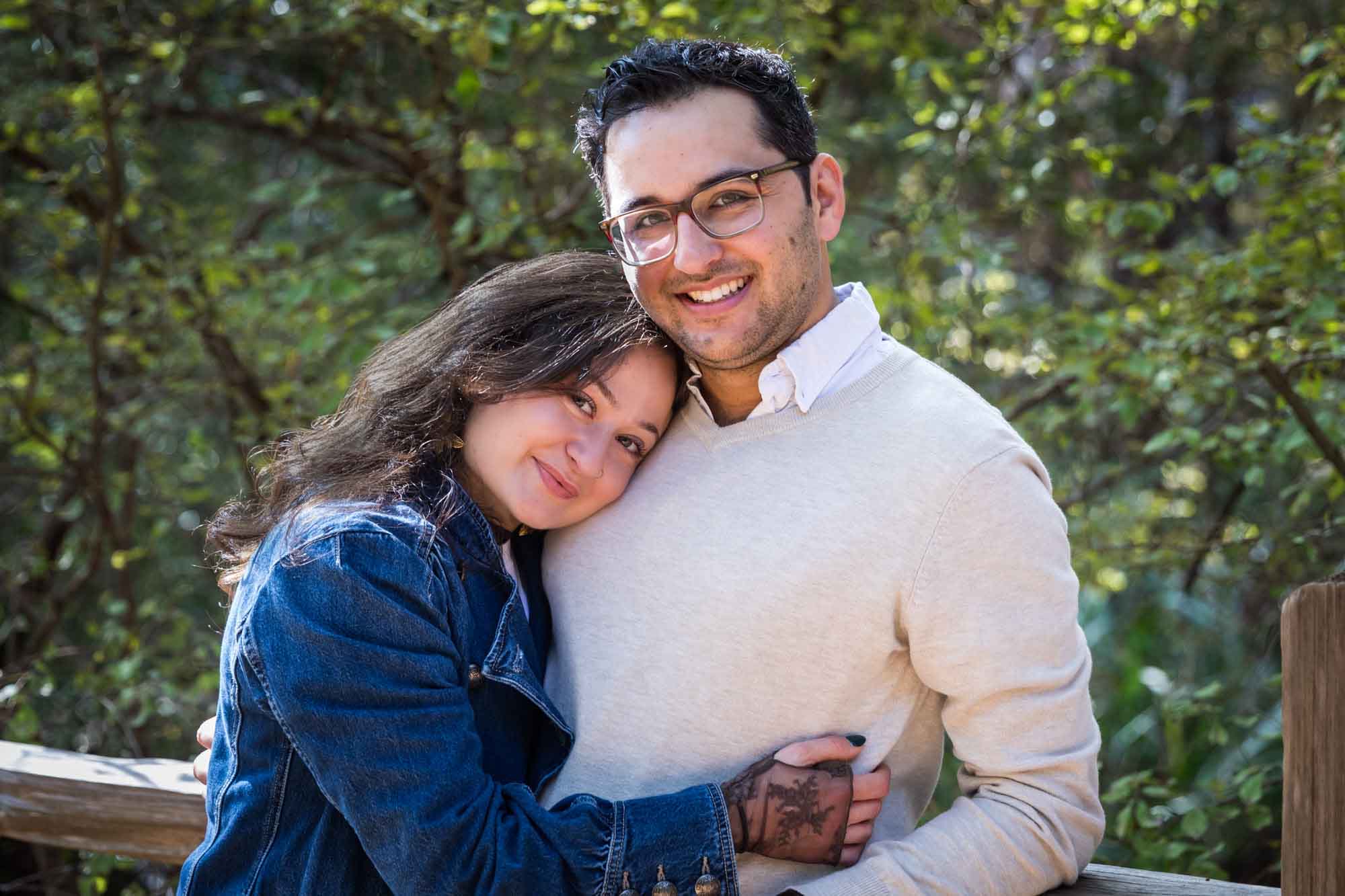 Woman wearing denim jacket hugging man wearing grey sweater against wooden railing in front of trees during a Red Bud Isle surprise proposal