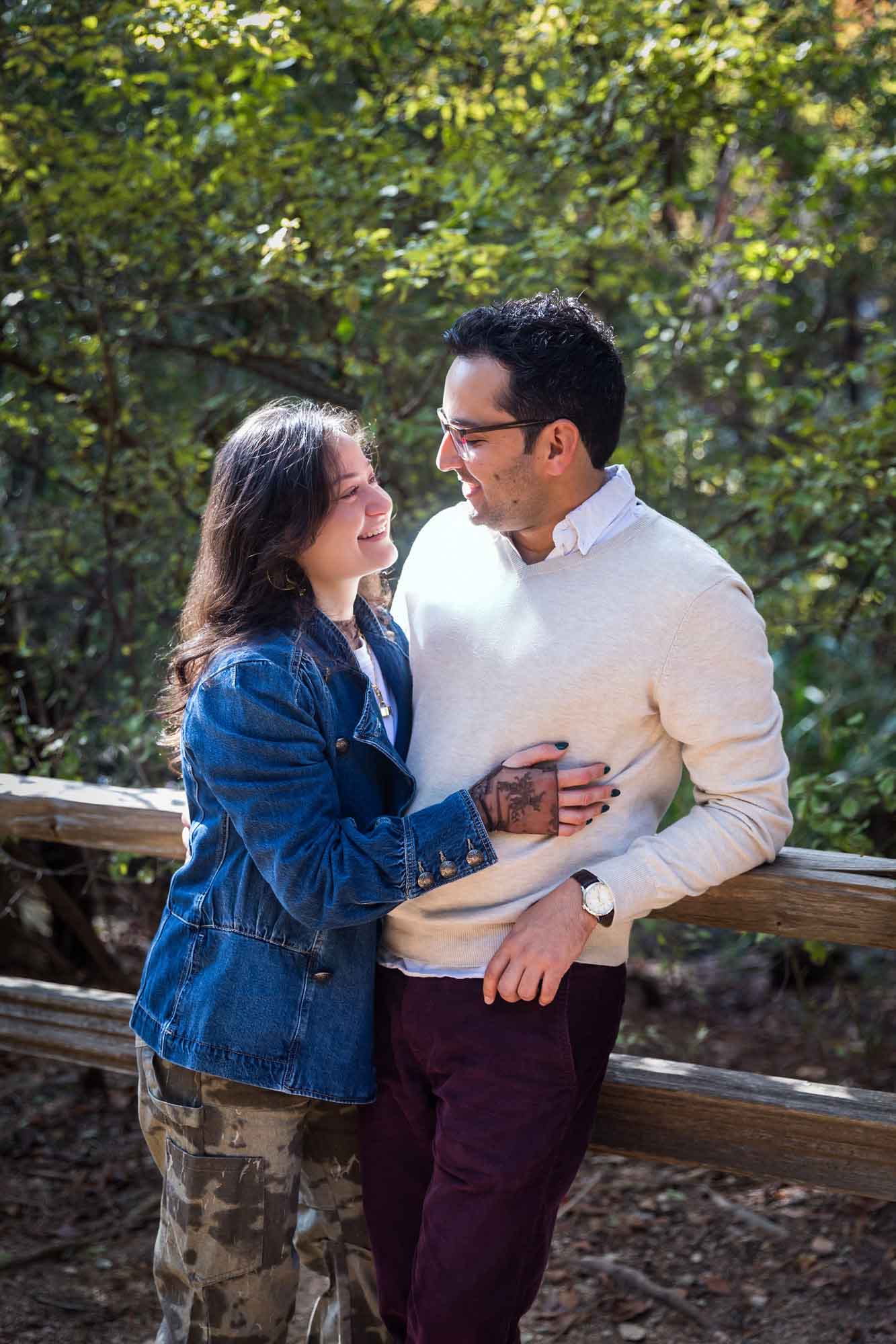 Woman wearing denim jacket hugging man wearing grey sweater against wooden railing in front of trees during a Red Bud Isle surprise proposal