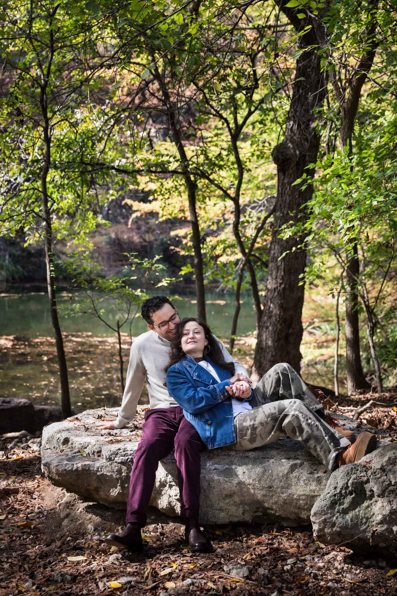 Woman wearing denim jacket leaning against man wearing grey sweater on a rock in the forest during a Red Bud Isle surprise proposal