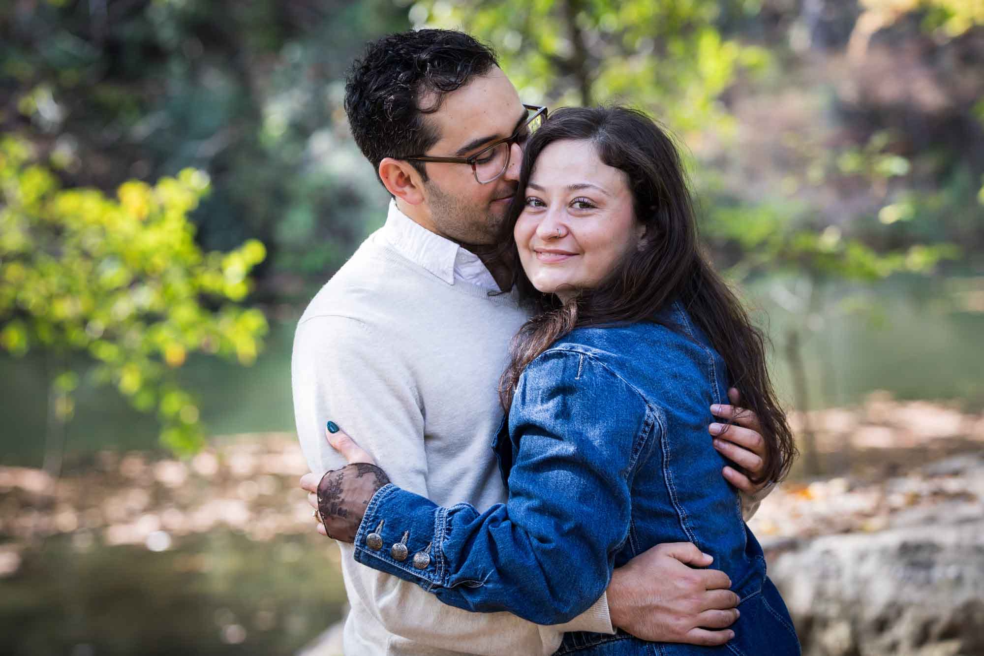 Woman wearing denim jacket hugging man wearing grey sweater in front of trees during a Red Bud Isle surprise proposal