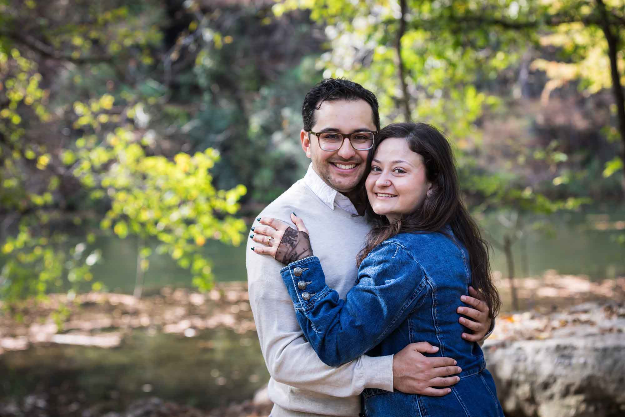 Woman wearing denim jacket hugging man wearing grey sweater in front of trees during a Red Bud Isle surprise proposal
