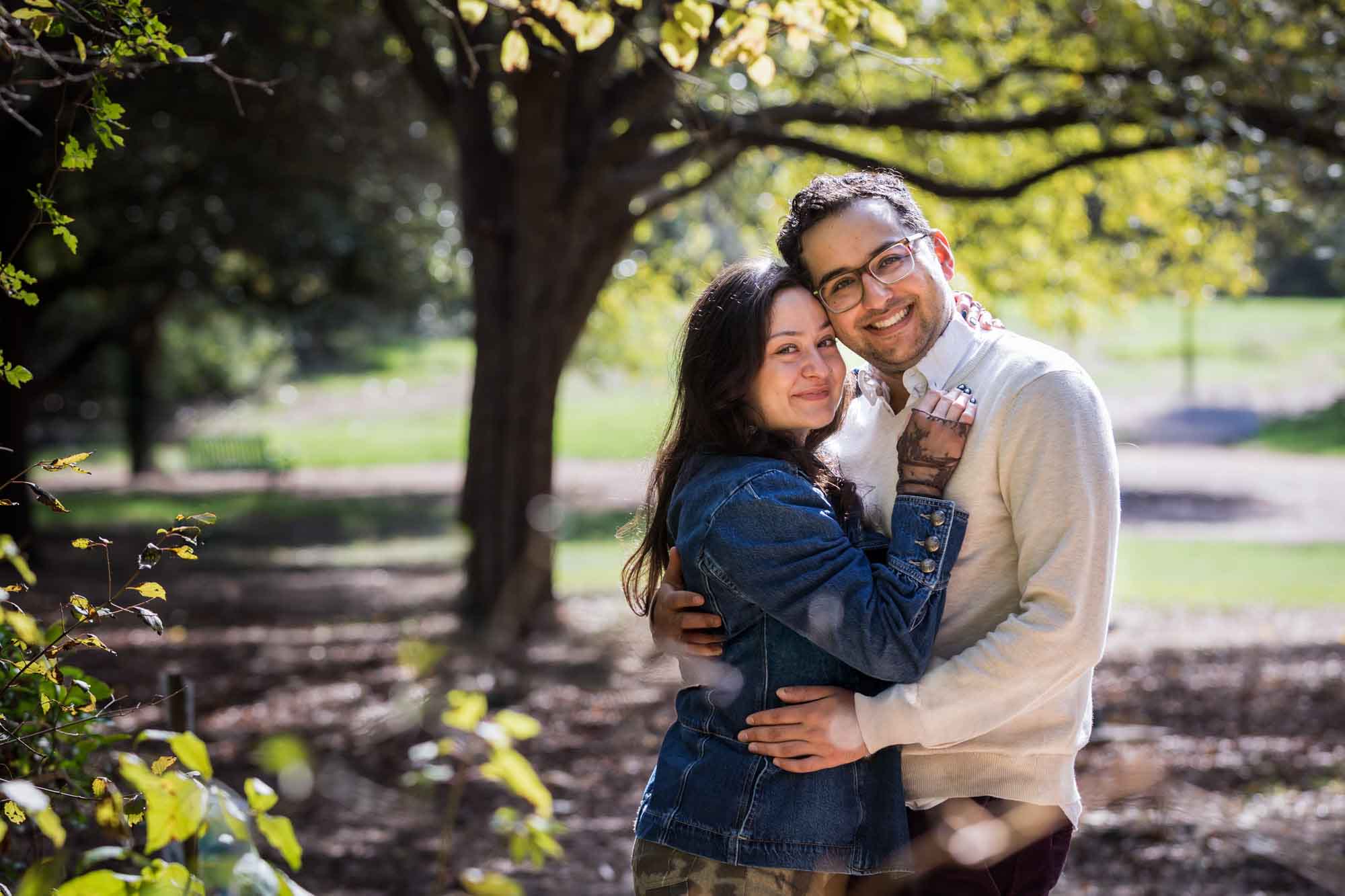 Woman wearing denim jacket hugging man wearing grey sweater in front of trees during a Red Bud Isle surprise proposal