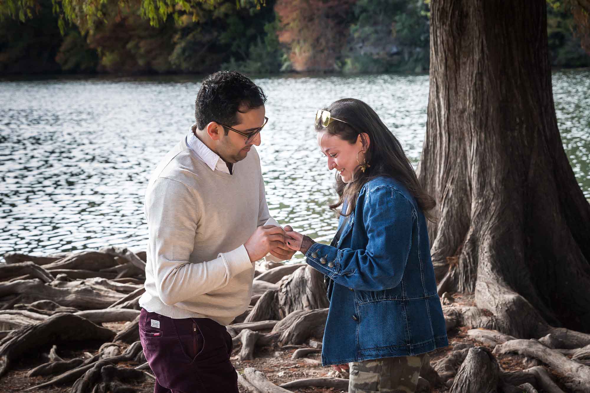 Man wearing maroon pants and white shirt putting ring on finger of woman wearing camo pants and denim jacket at end of island with tree in background during a Red Bud Isle surprise proposal