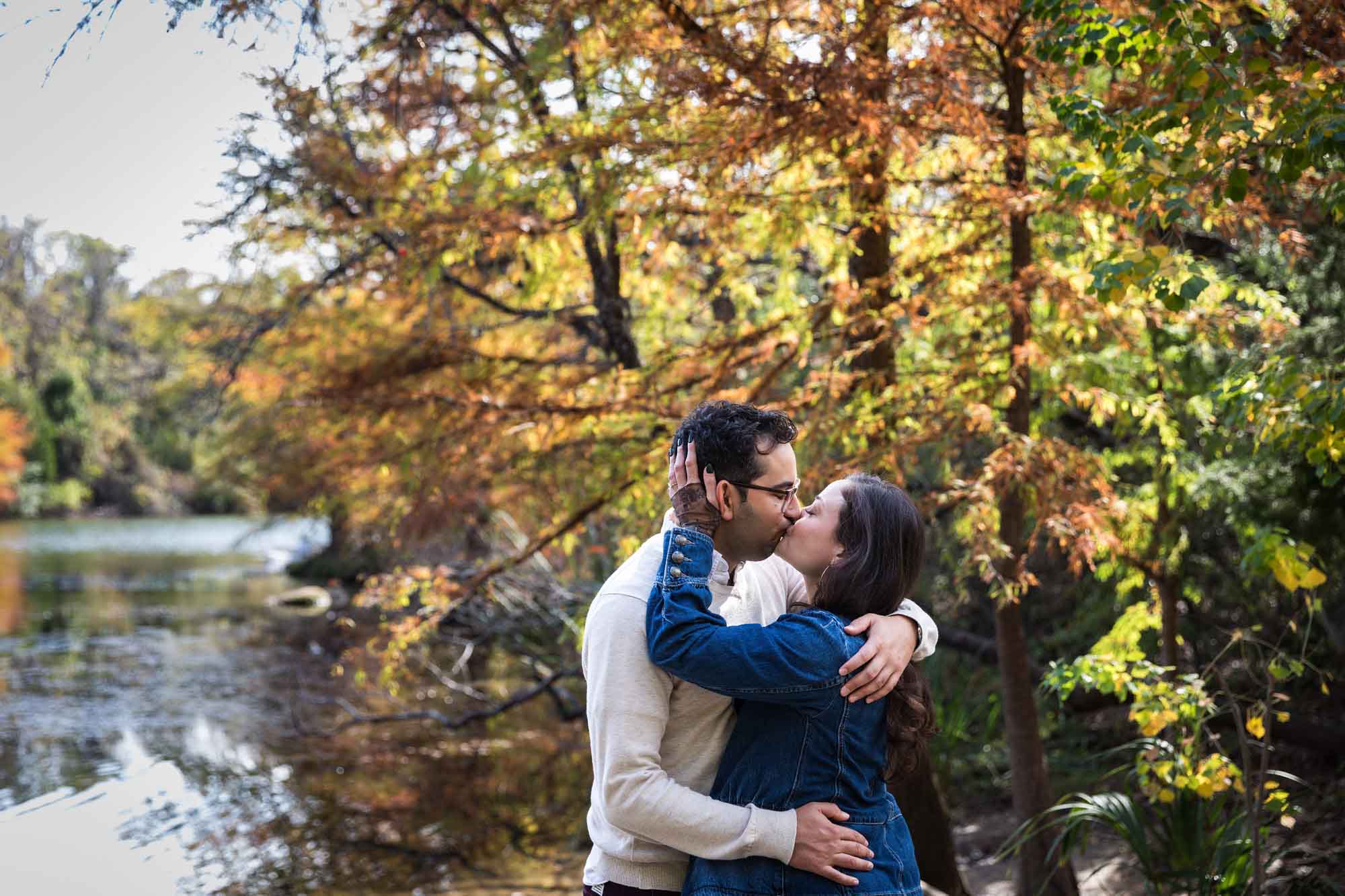 Woman wearing denim jacket kissing man wearing grey sweater in front of colorful trees and river during a Red Bud Isle surprise proposal