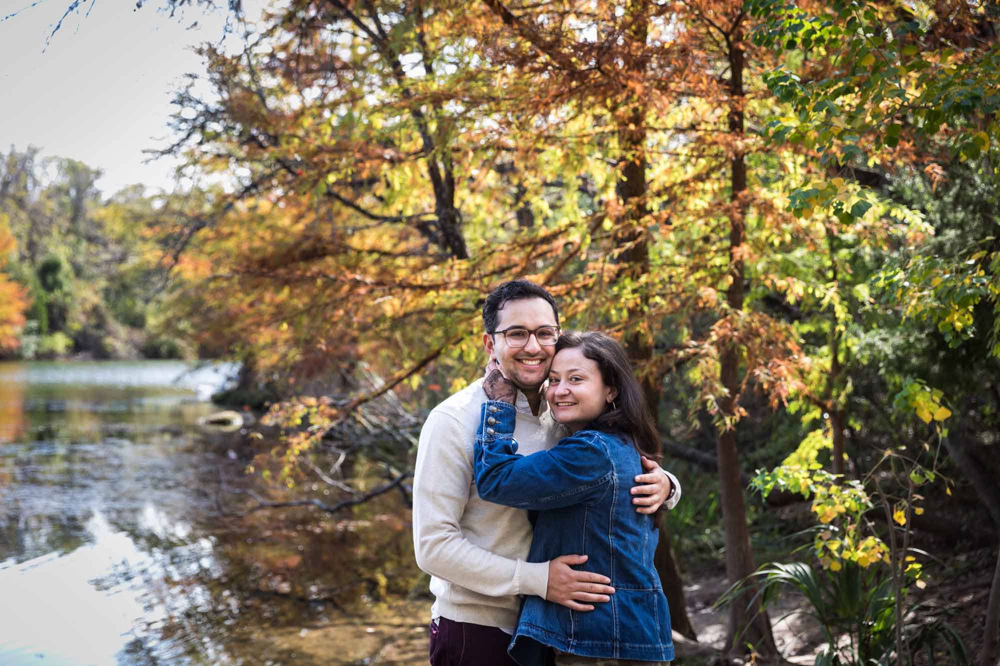 Woman wearing denim jacket hugging man wearing grey sweater in front of colorful trees and river during a Red Bud Isle surprise proposal