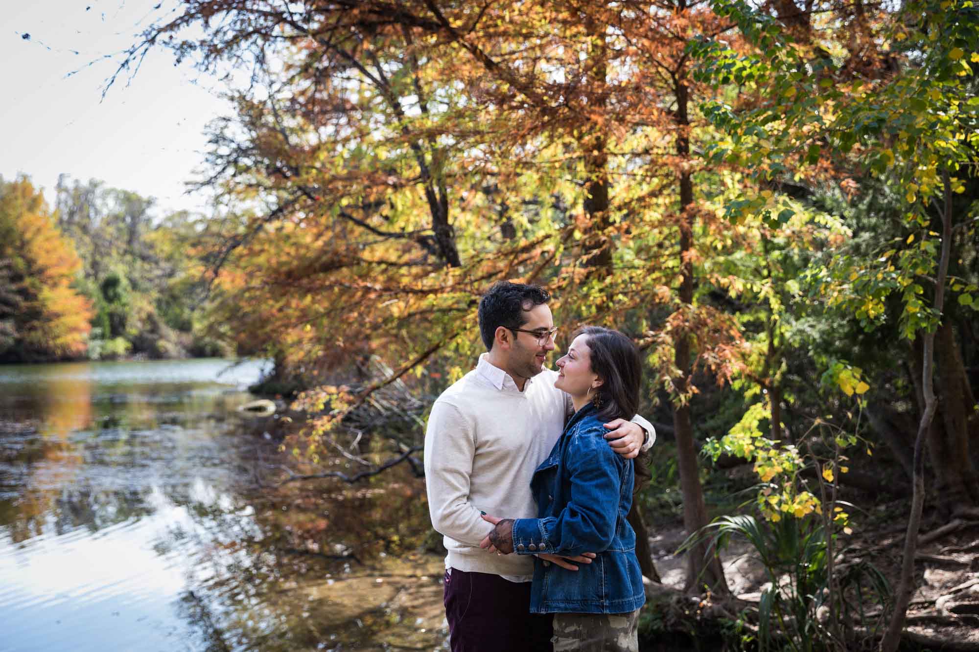 Woman wearing denim jacket hugging man wearing grey sweater in front of colorful trees and river during a Red Bud Isle surprise proposal