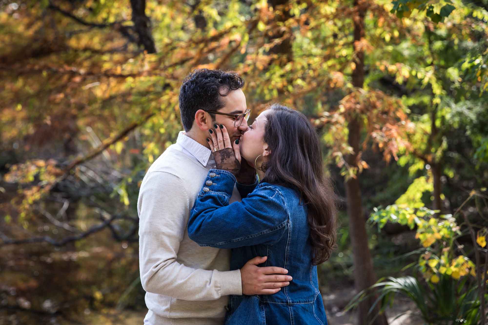 Woman wearing denim jacket kissing man wearing grey sweater in front of colorful trees and river during a Red Bud Isle surprise proposal