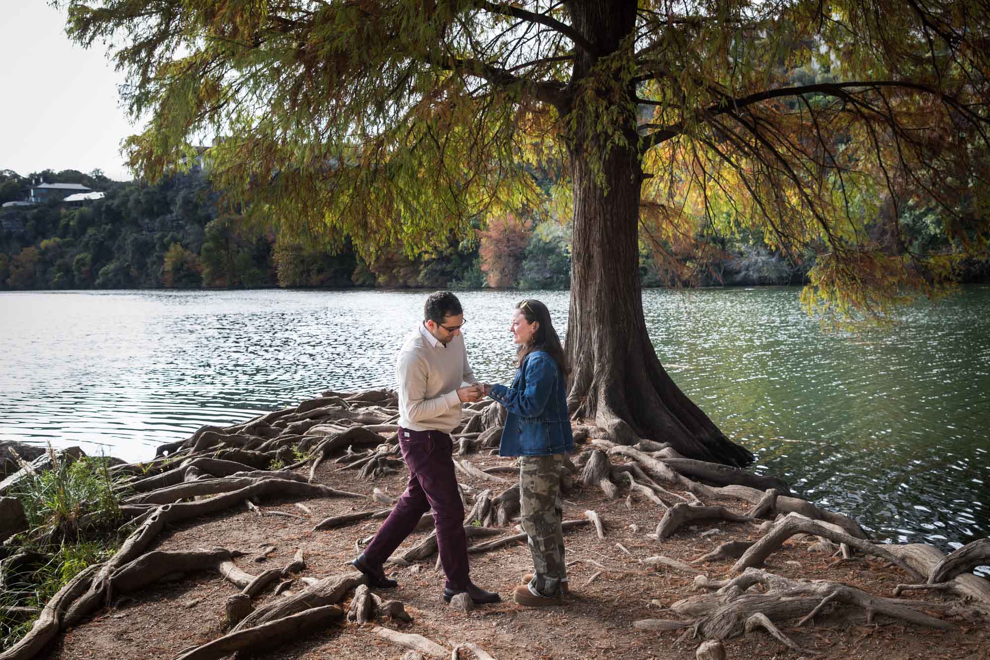 Man wearing maroon pants and white shirt putting ring on finger of woman wearing camo pants and denim jacket at end of island with tree in background during a Red Bud Isle surprise proposal