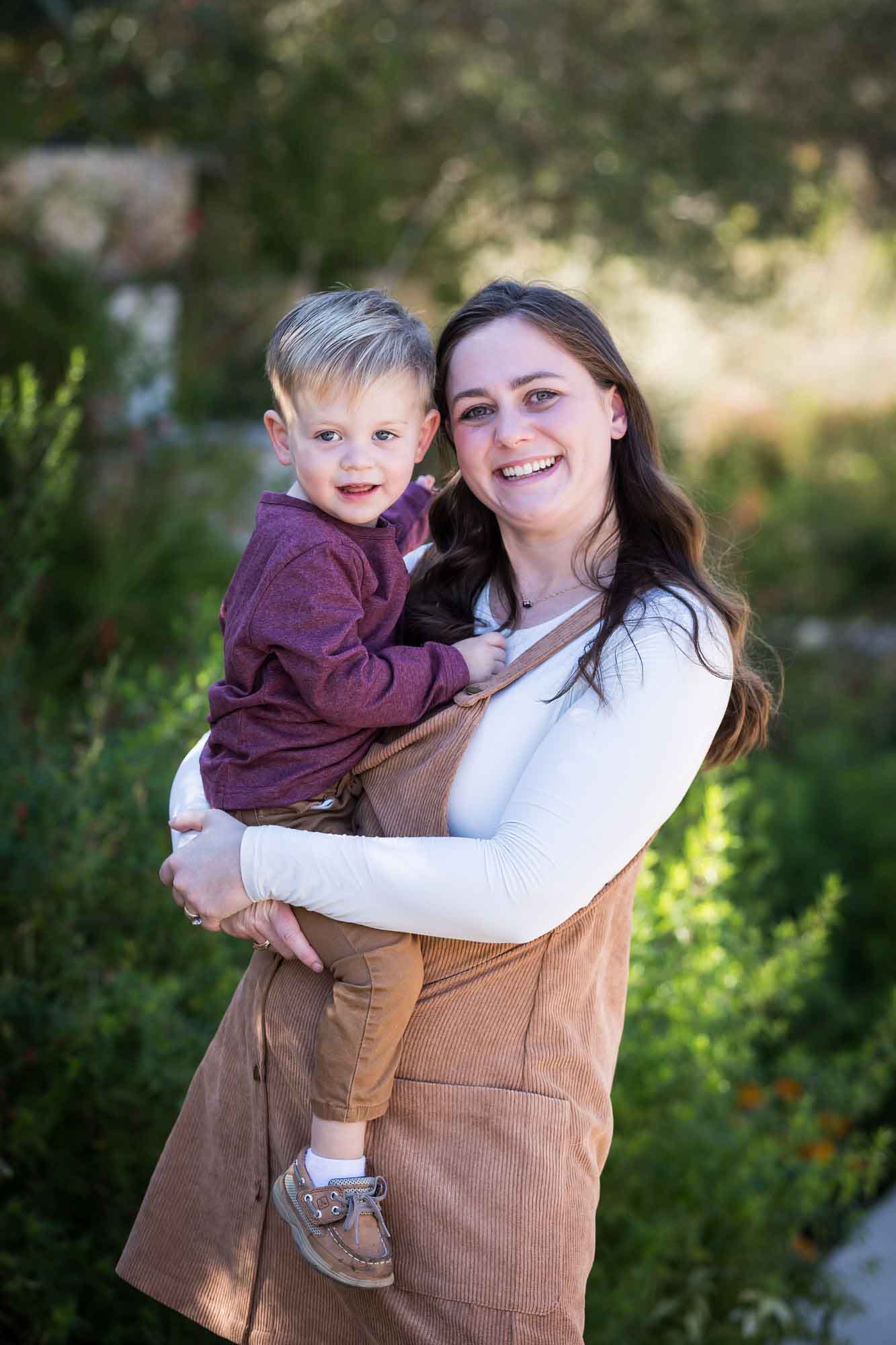 San Antonio Botanical Gardens family portrait of woman wearing white shirt and corduroy dress holding little boy wearing purple shirt and tan pants