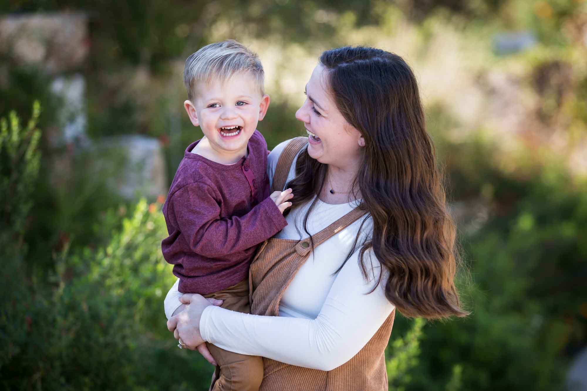 San Antonio Botanical Gardens family portrait of woman wearing white shirt and corduroy dress holding little boy wearing purple shirt and tan pants