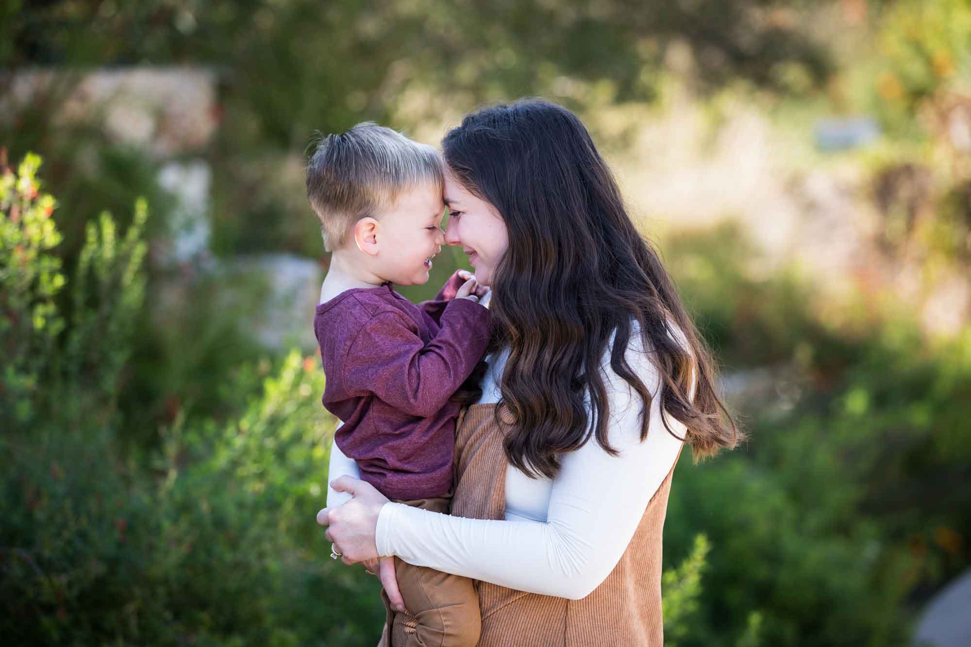 San Antonio Botanical Gardens family portrait of woman wearing white shirt and corduroy dress holding little boy wearing purple shirt and tan pants and touching foreheads