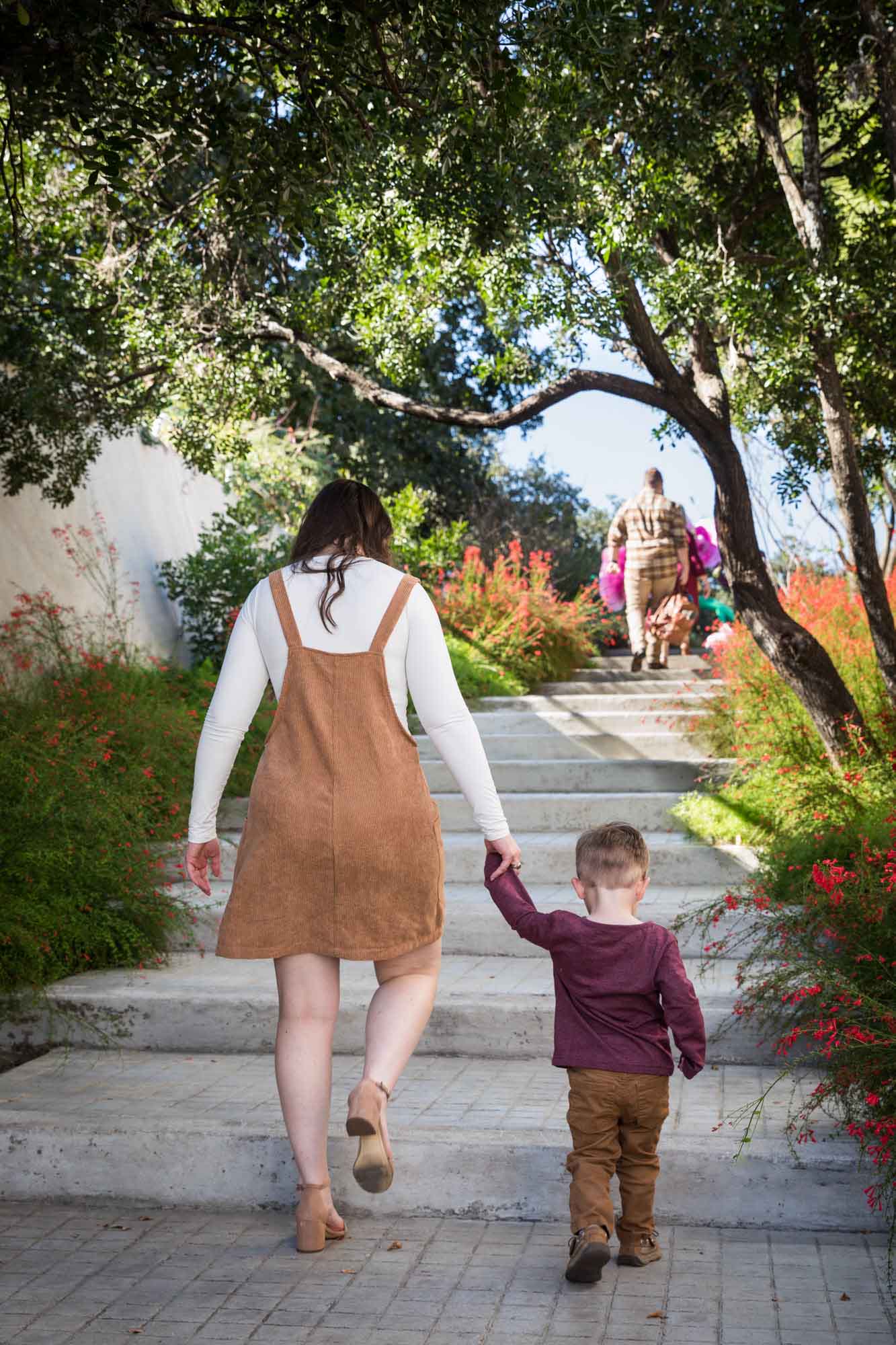 Mother wearing corduroy dress holding hand of little boy wearing maroon shirt walking up stairs during a San Antonio Botanical Gardens family portrait session
