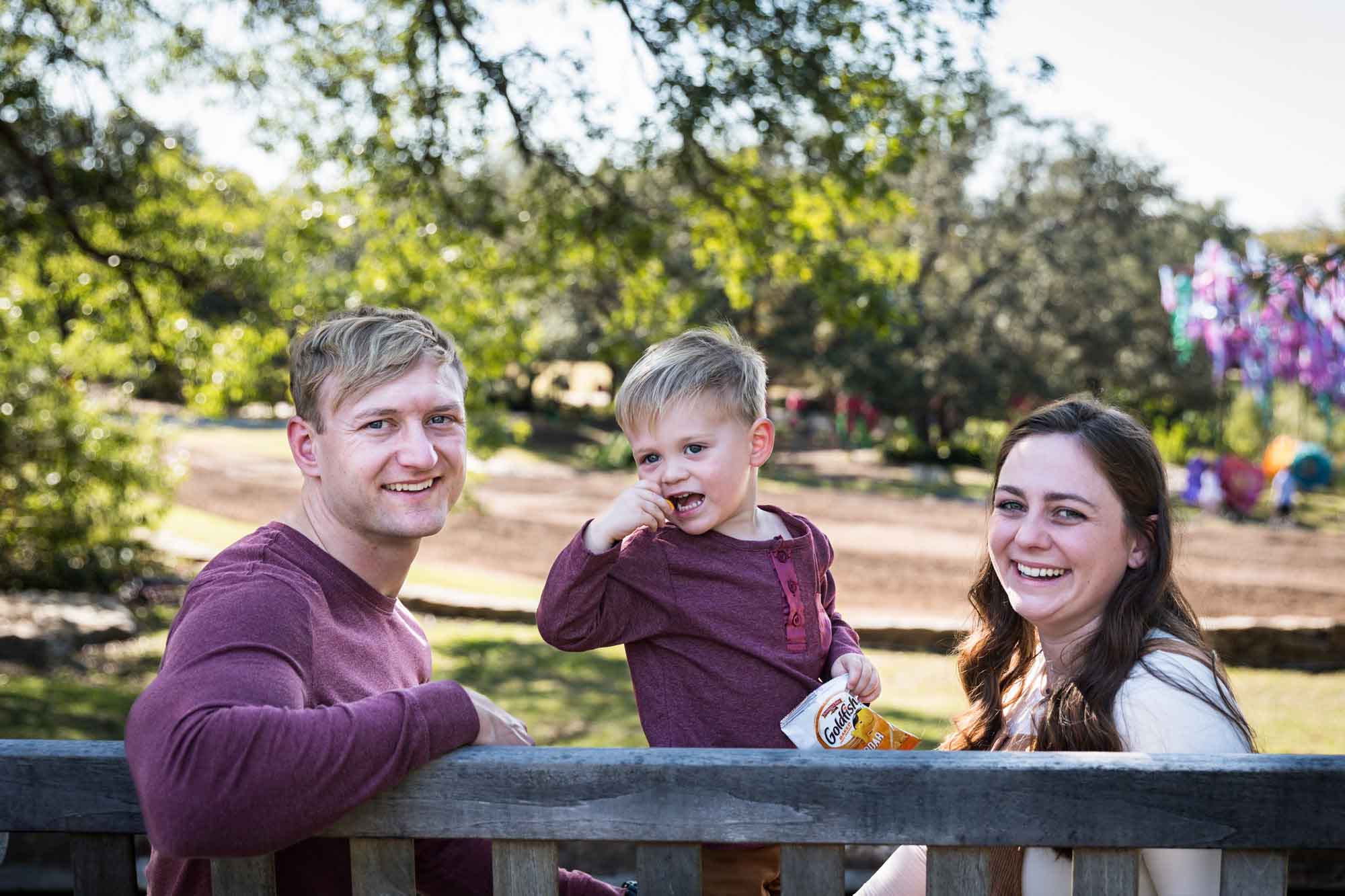 San Antonio Botanical Gardens family portrait of mother, father, and little boy who is eating goldfish crackers on a wooden bench