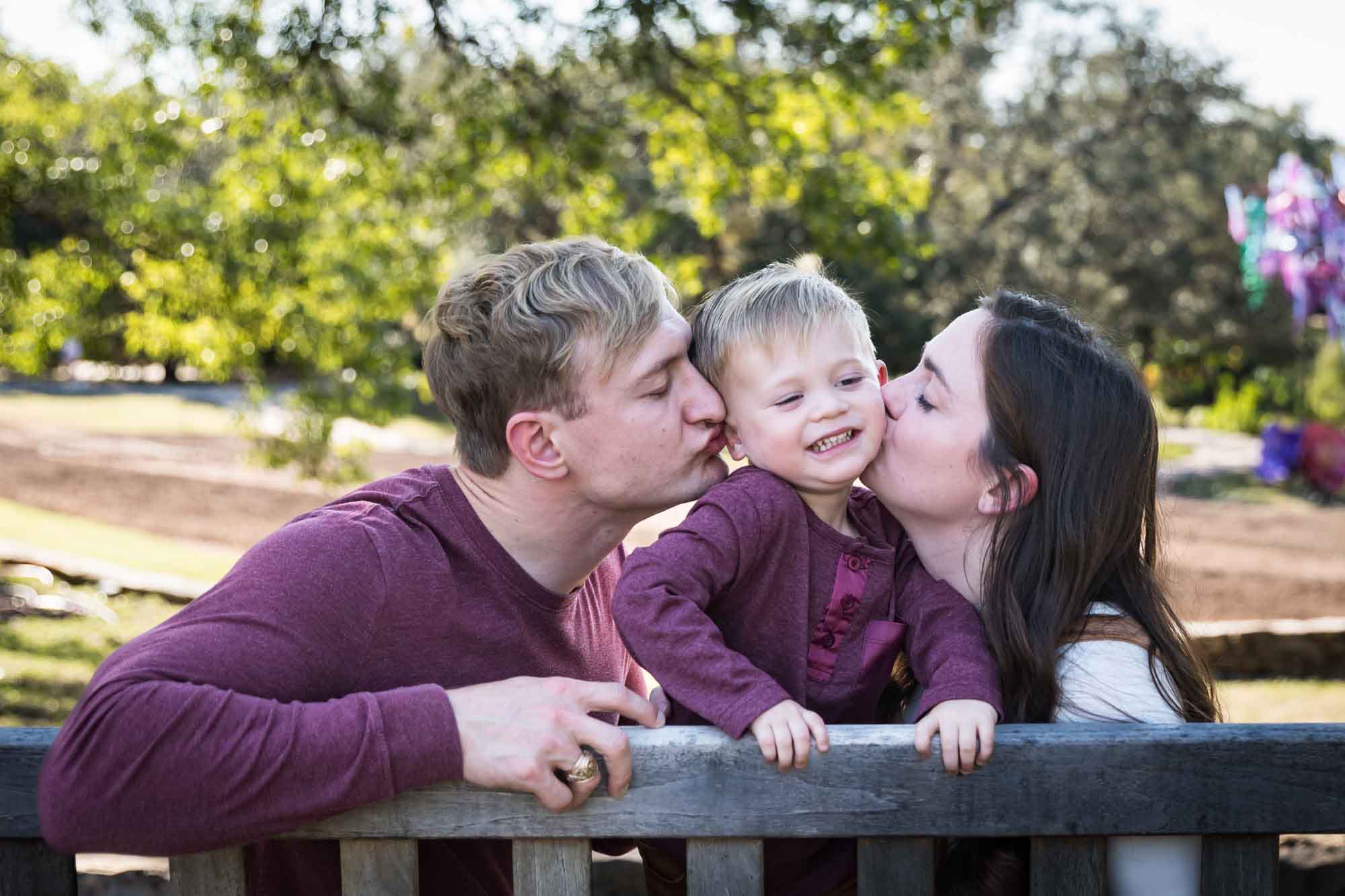 San Antonio Botanical Gardens family portrait of mother and father kissing little boy on the cheeks on a wooden bench