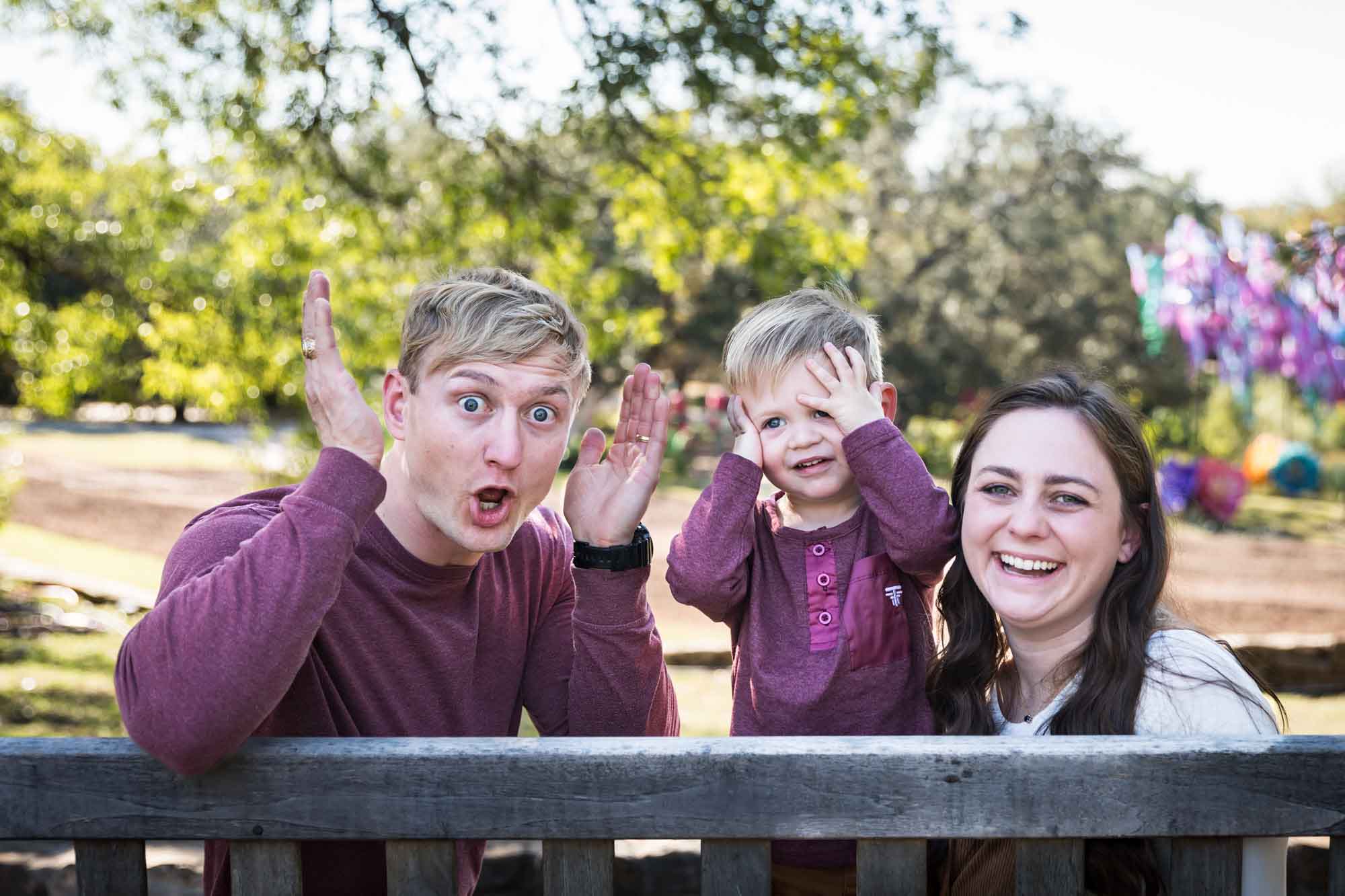 San Antonio Botanical Gardens family portrait of mother and father playing peekaboo with little boy on wooden bench