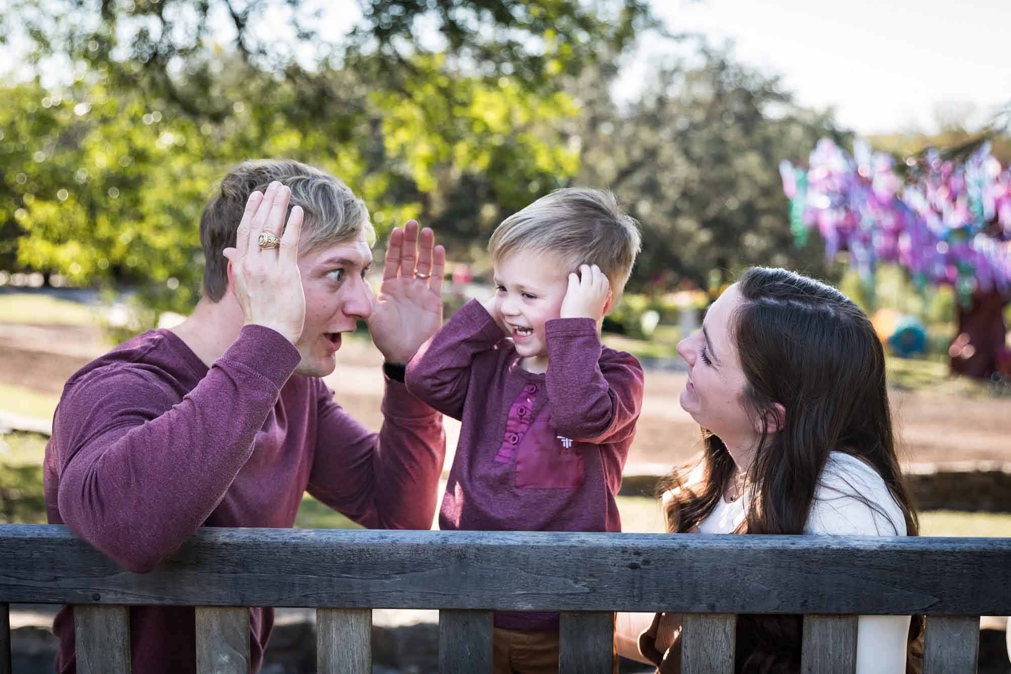 San Antonio Botanical Gardens family portrait of mother and father playing peekaboo with little boy on wooden bench